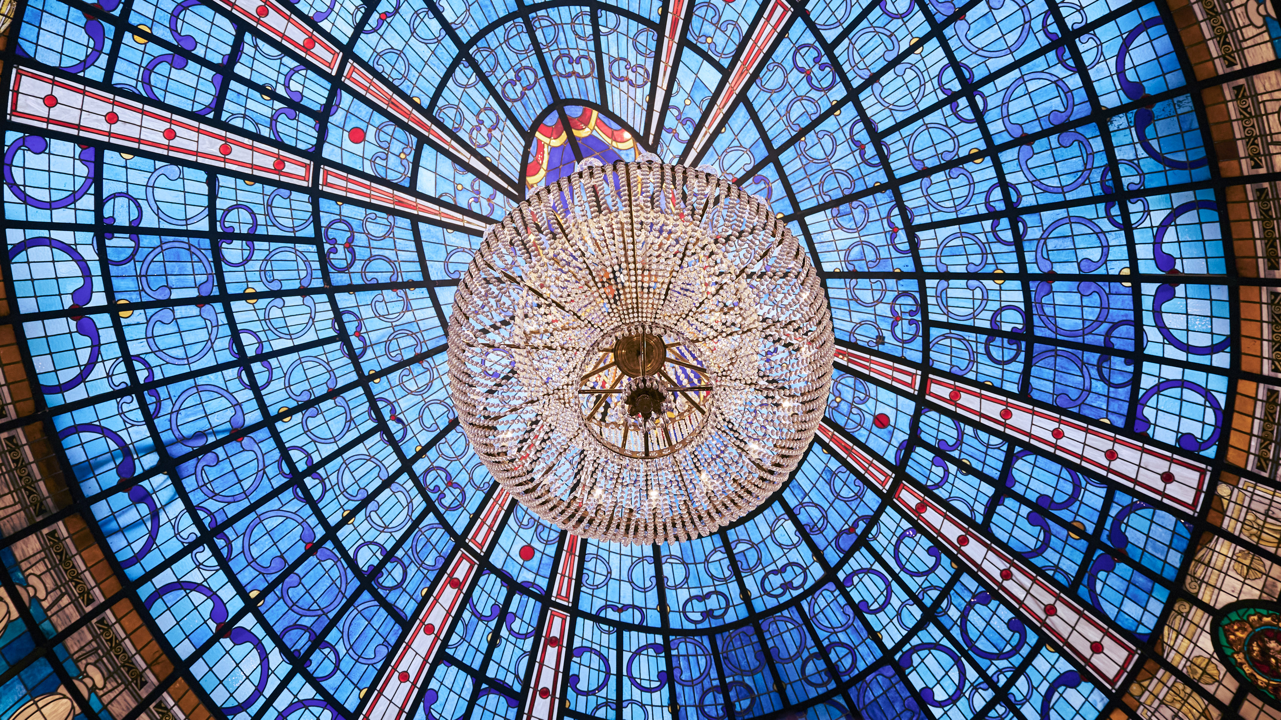 Ornate crystal chandelier under an intricate blue stained glass ceiling dome.