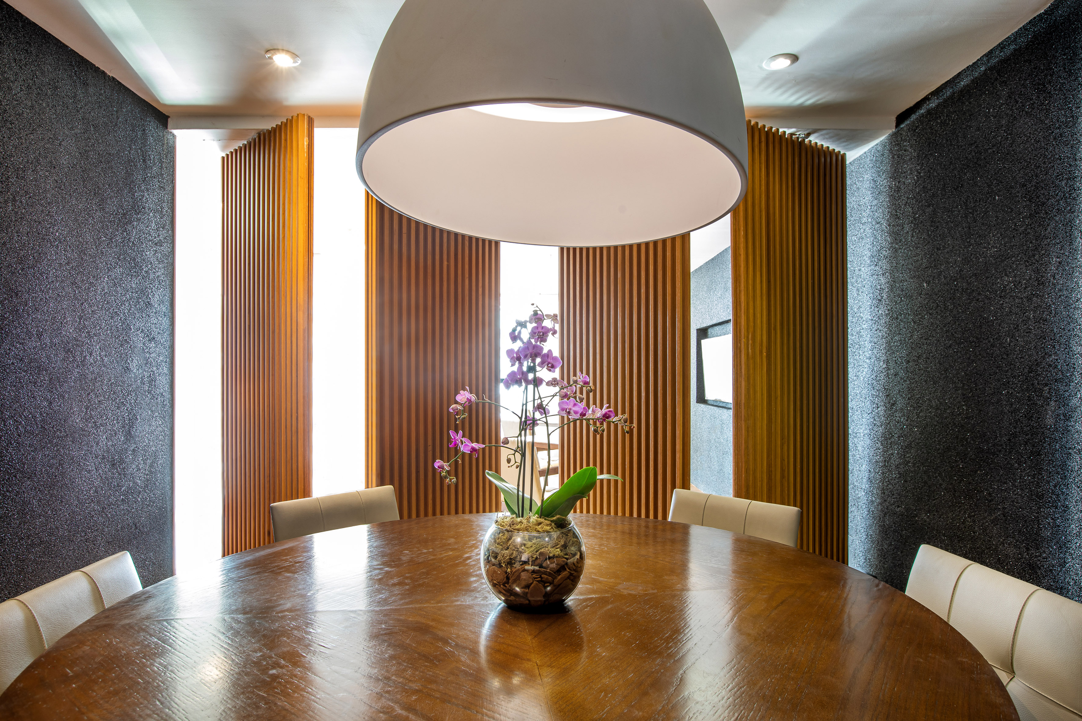 Modern dining room: round wood table, purple orchid, dark wall, wood panels.