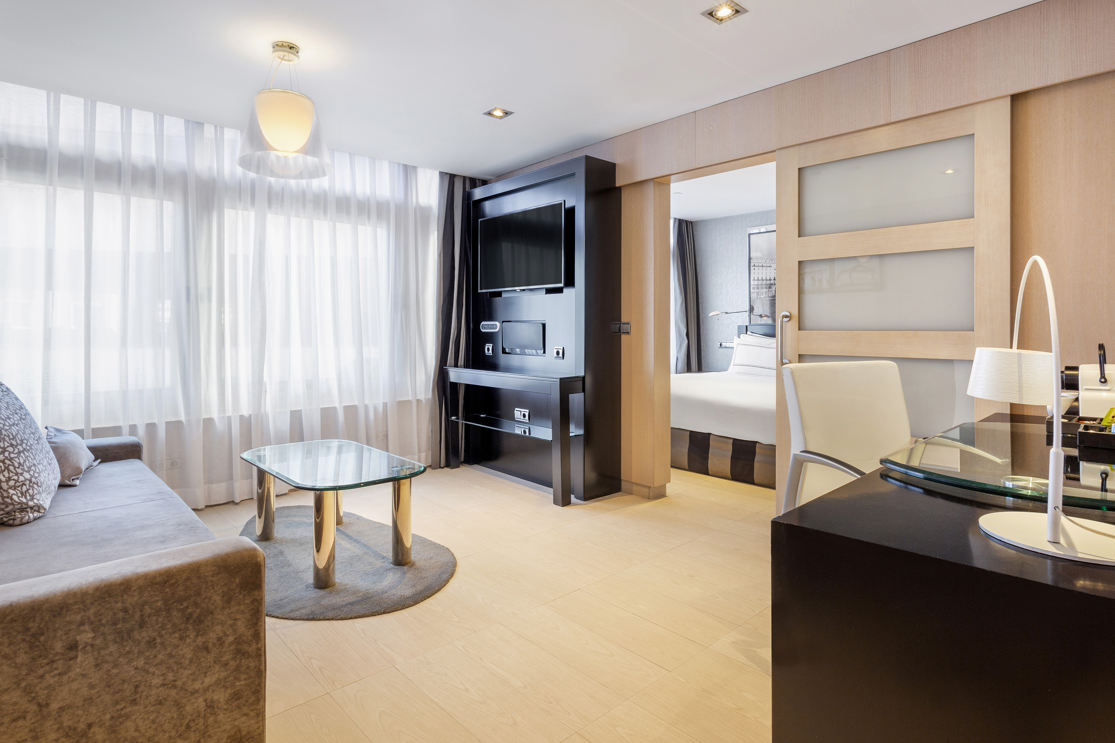 Modern hotel suite: living area, bedroom, desk, TV, light wood floor.