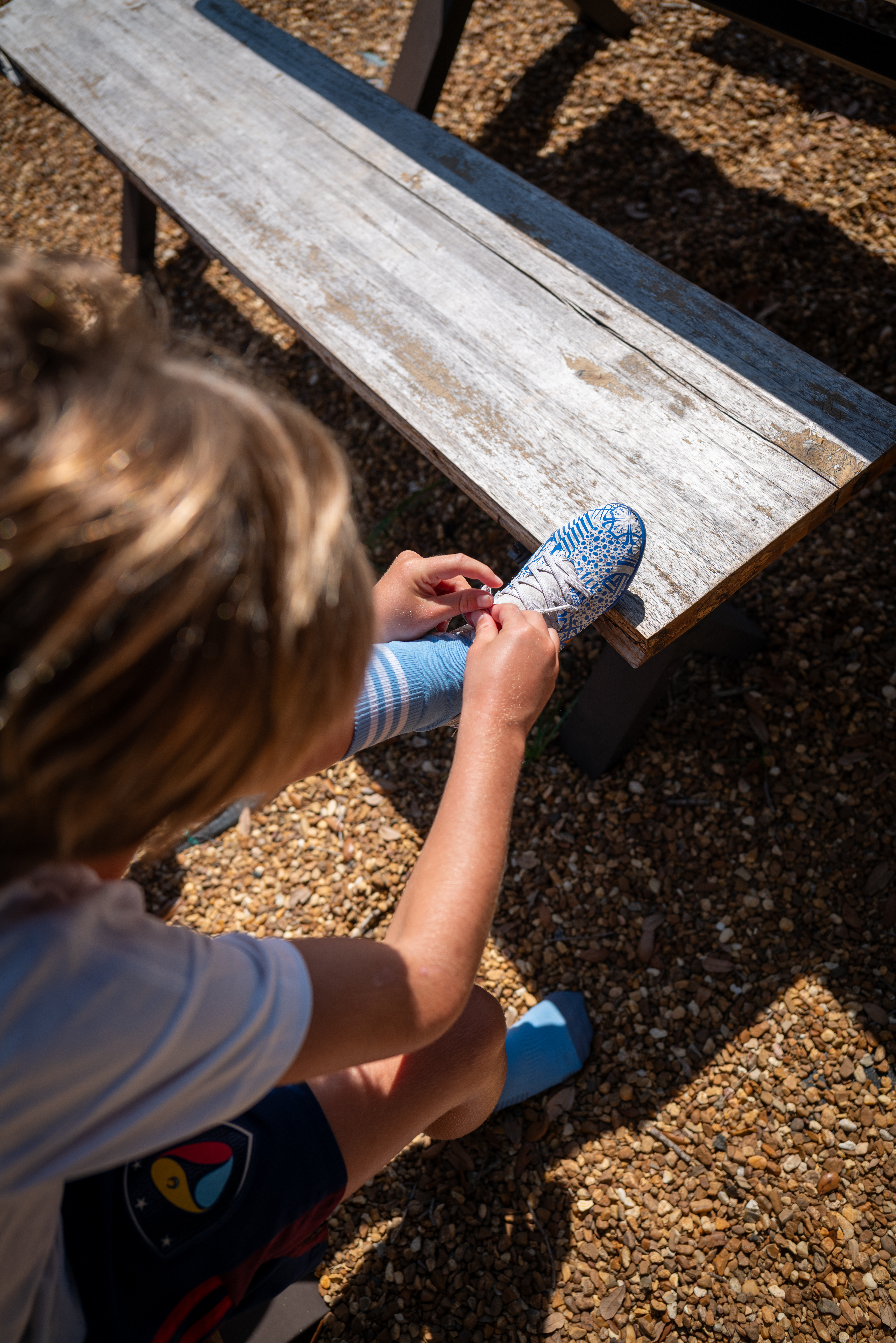 Child tying laces on blue patterned sports shoe outdoors, on gravel next to a bench.
