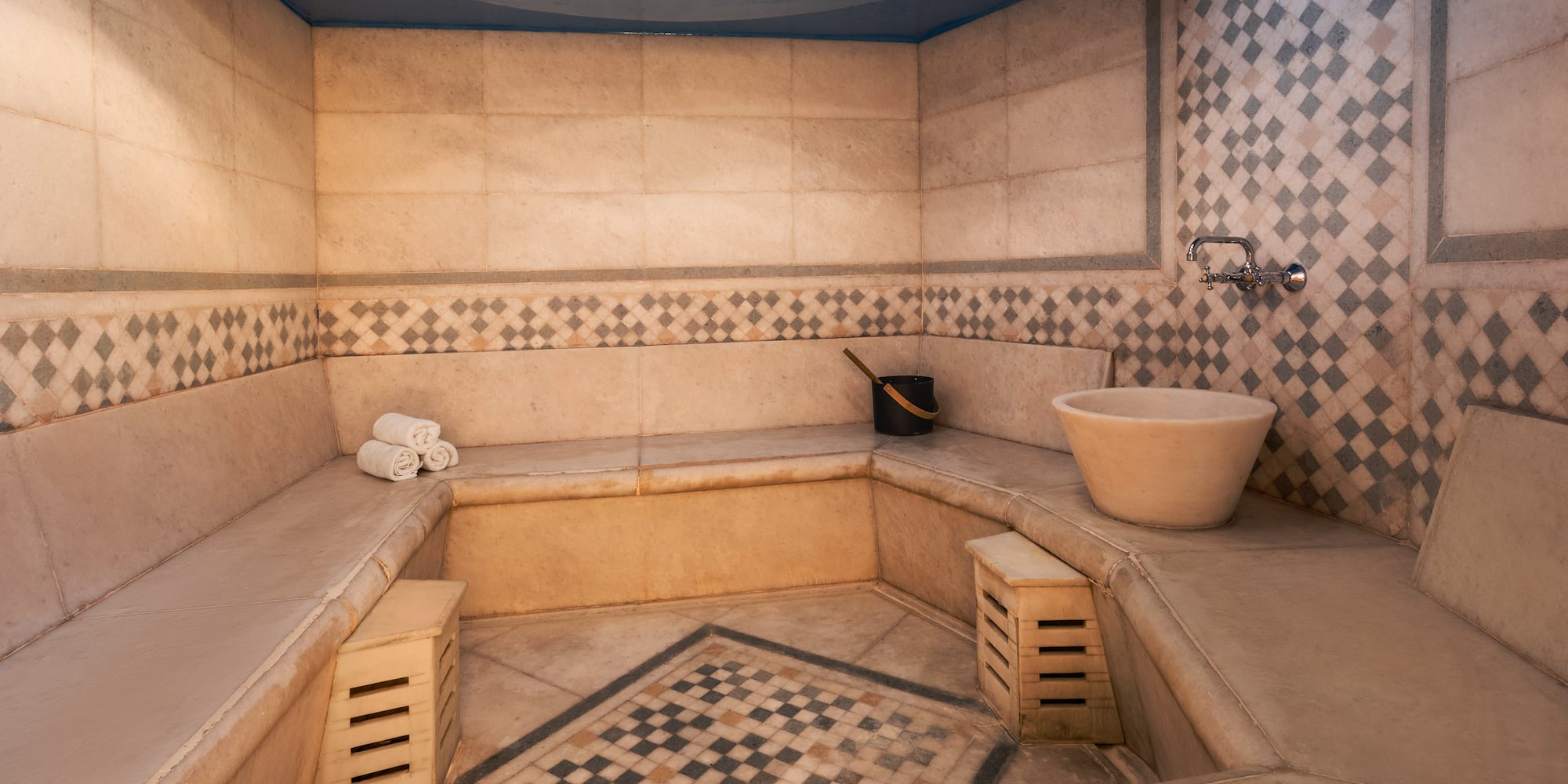 Tiled hammam with stone benches, basin, blue ceiling, and geometric patterns.