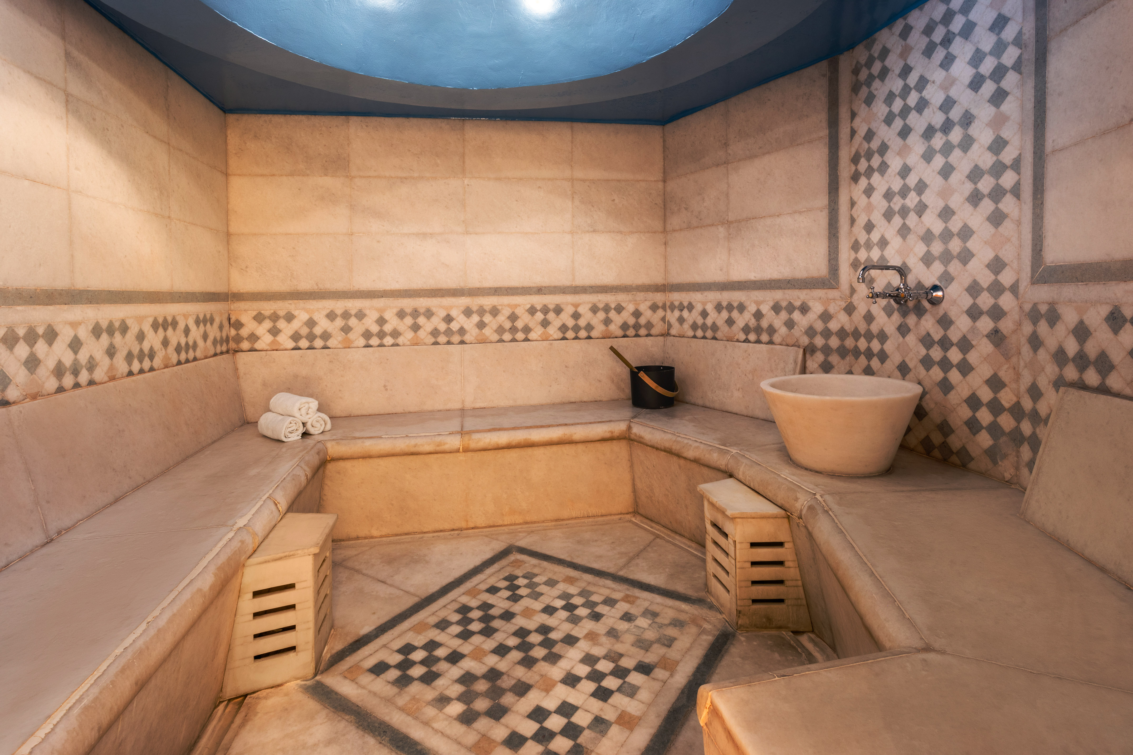 Tiled hammam with stone benches, basin, blue ceiling, and geometric patterns.