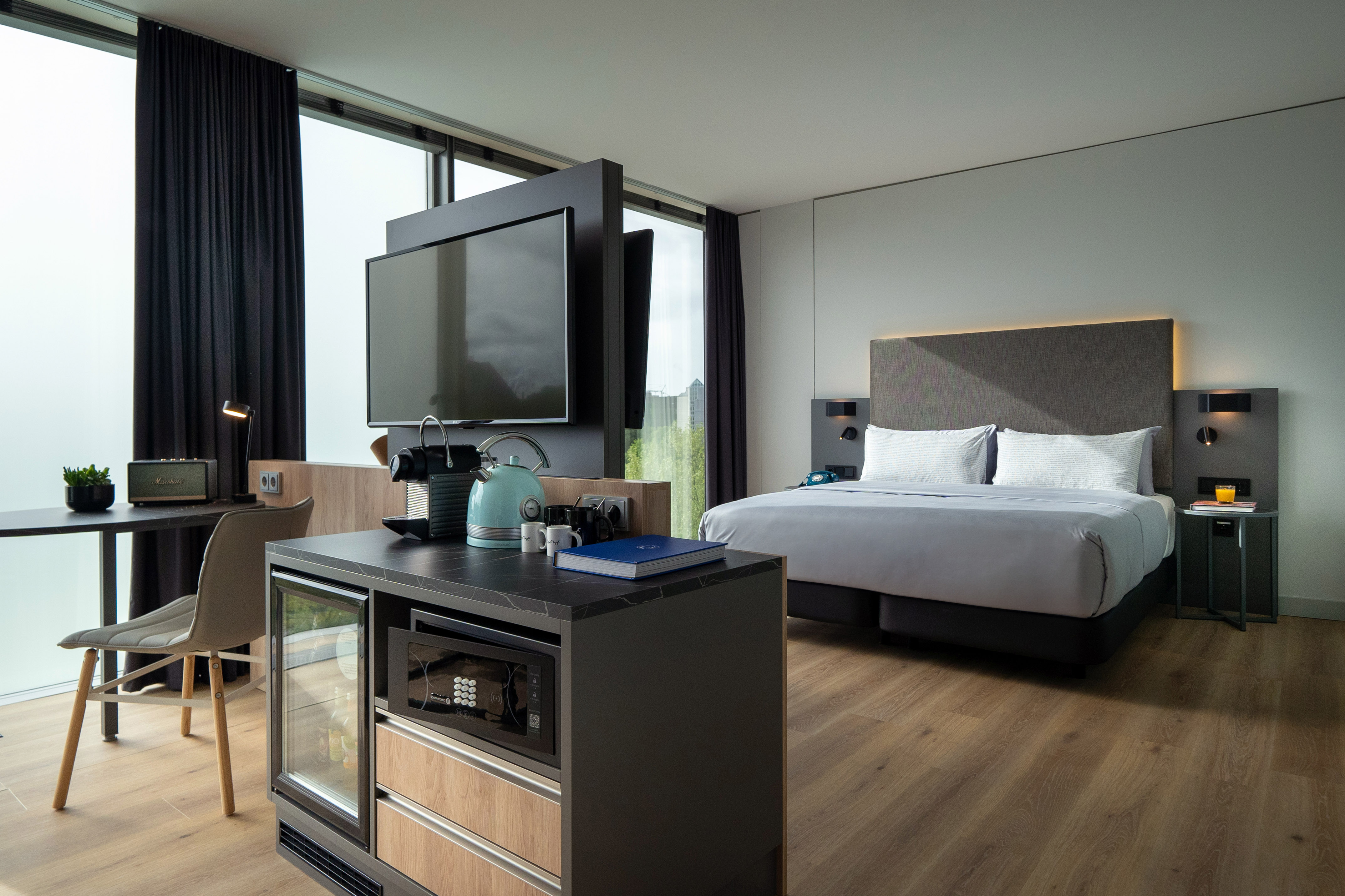 Modern hotel room with king bed, TV, minibar, and large panoramic windows.