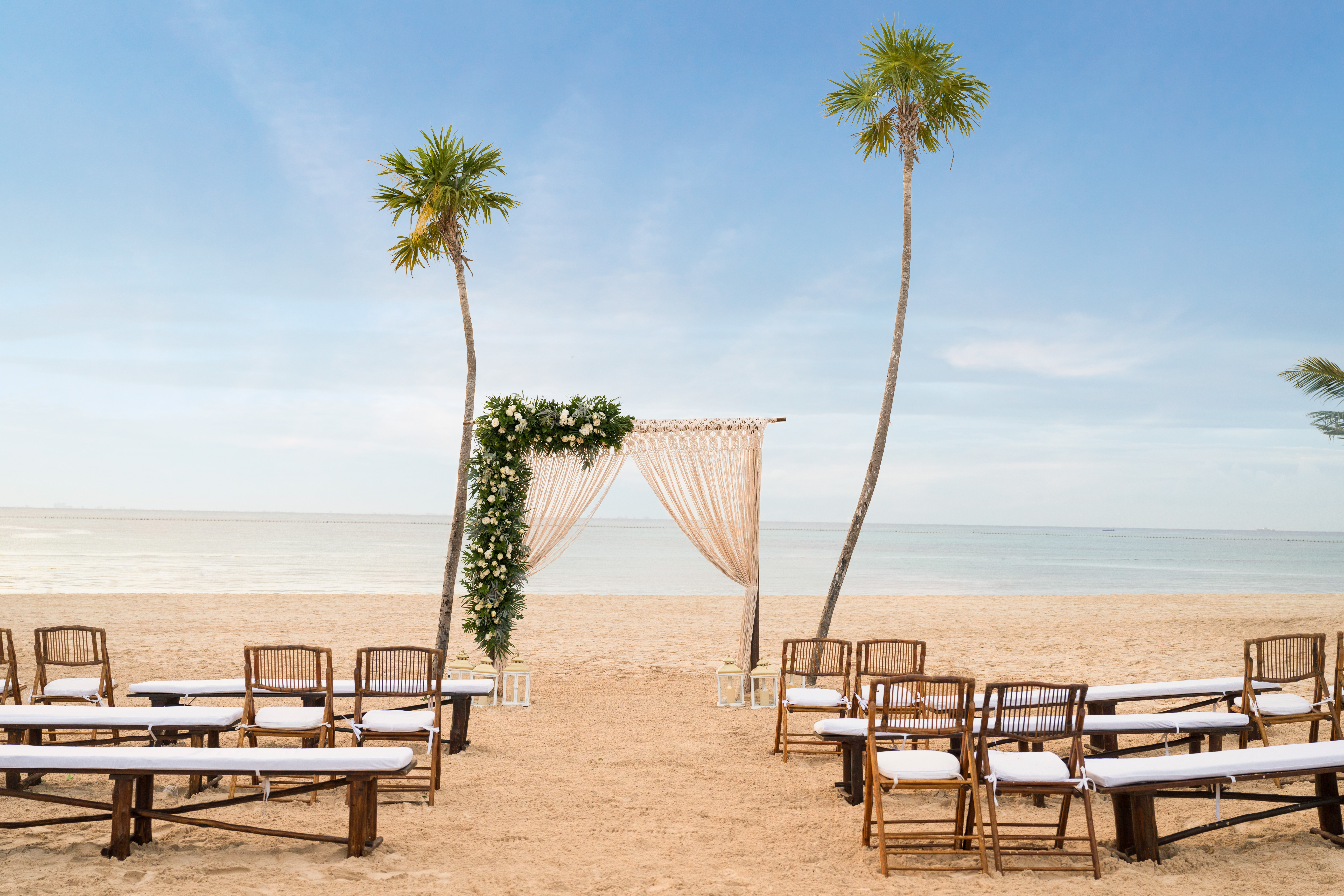 Tropical beach wedding setup with floral arch, chairs, and palm trees.
