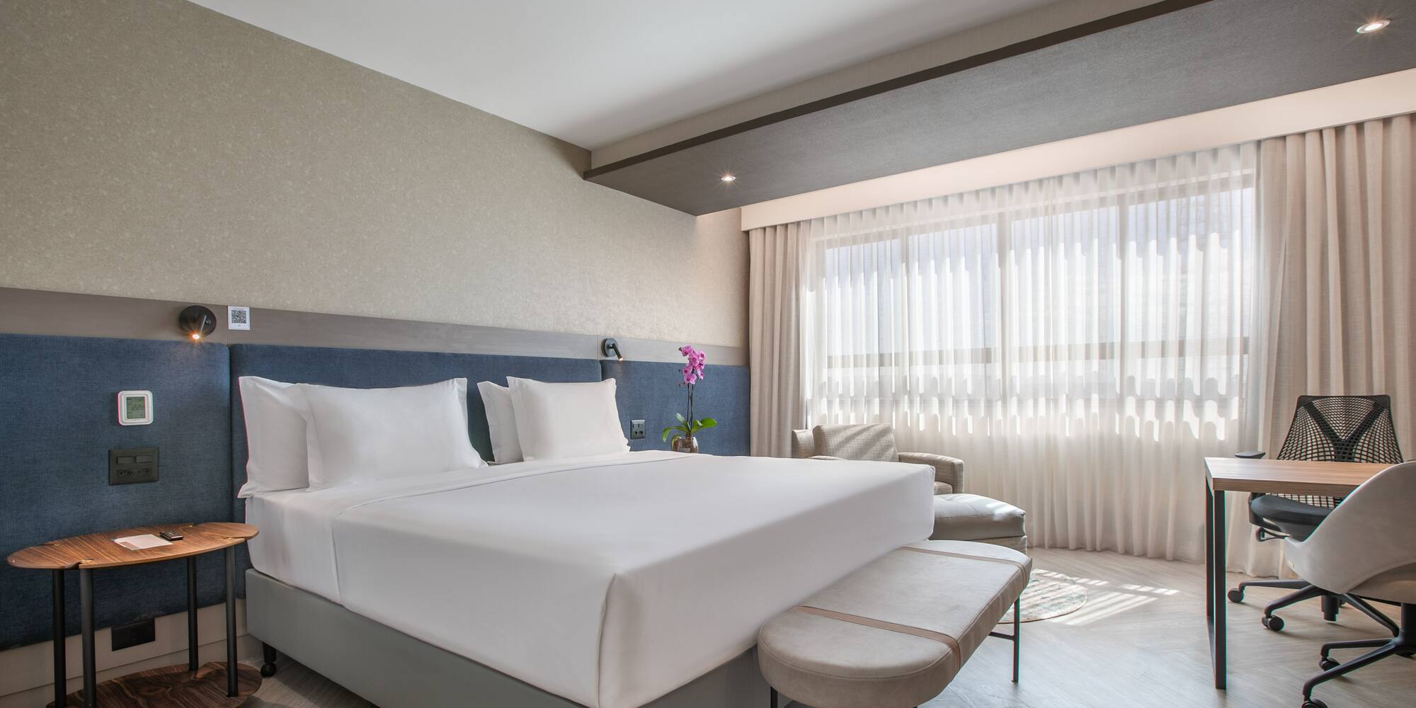 Clean, modern hotel room with king bed, blue headboard, desk, and bright window.