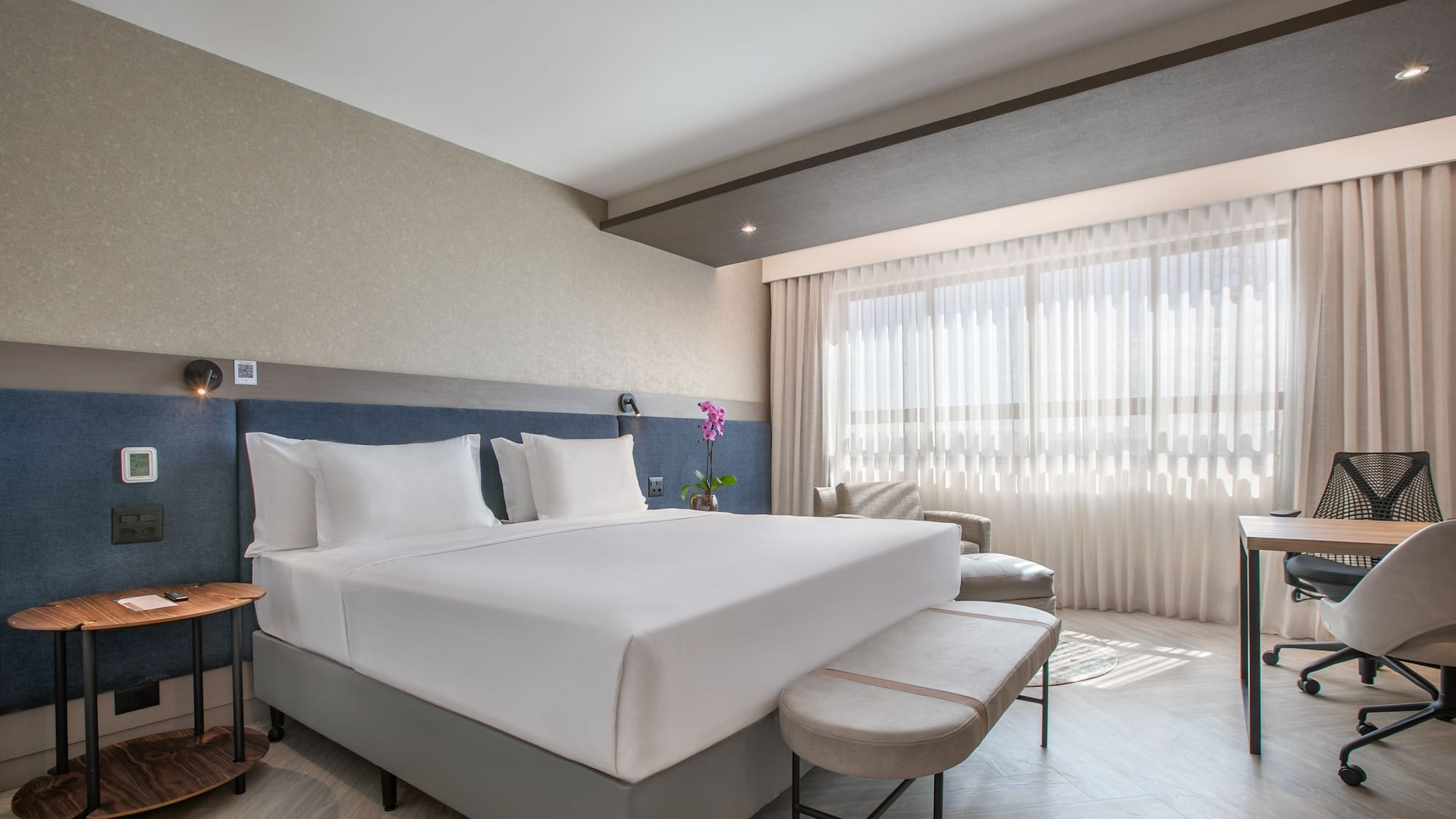 Contemporary hotel bedroom suite with king bed, desk area, and natural light.