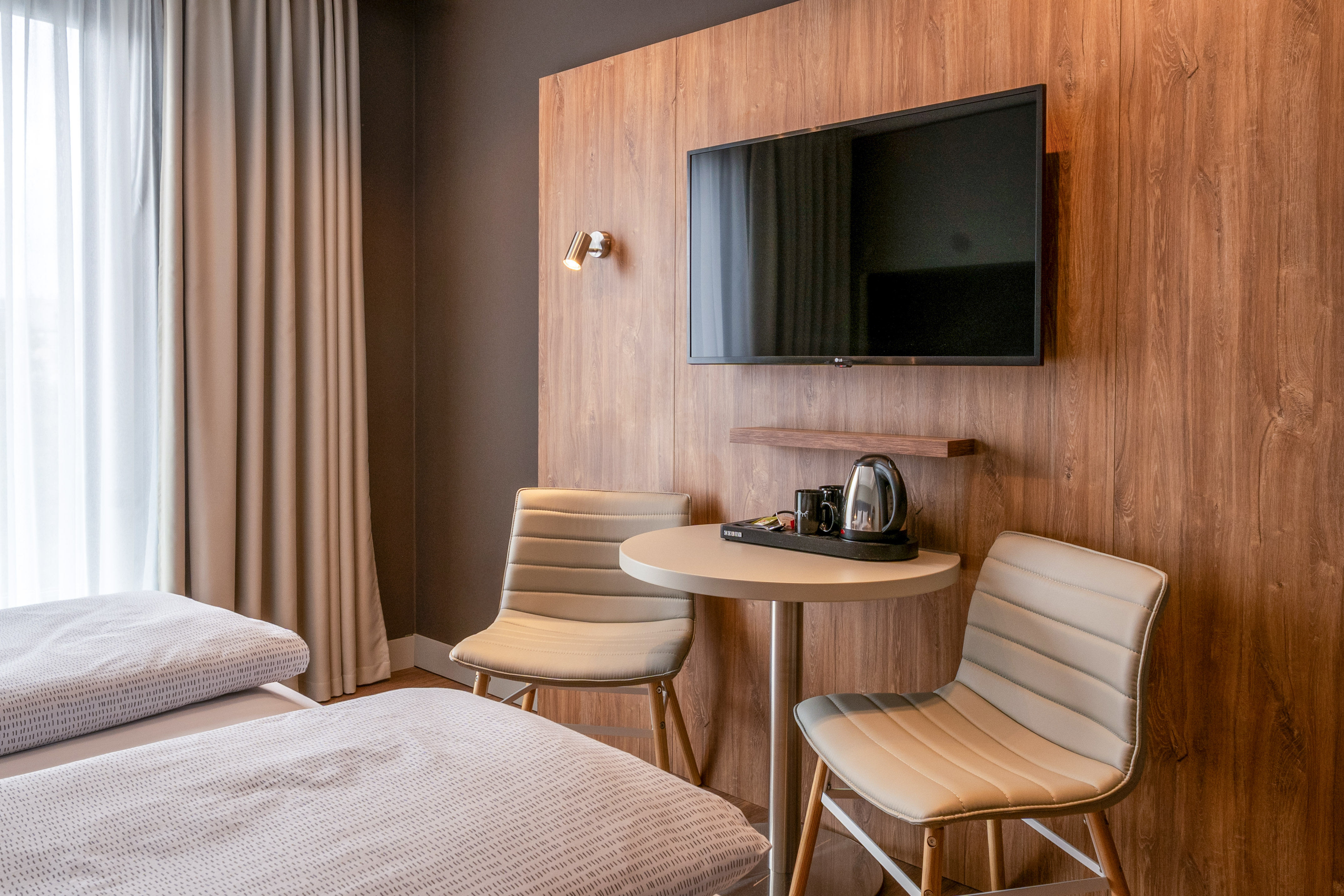 Modern hotel room with twin beds, wall TV, and coffee/tea station on wood wall.