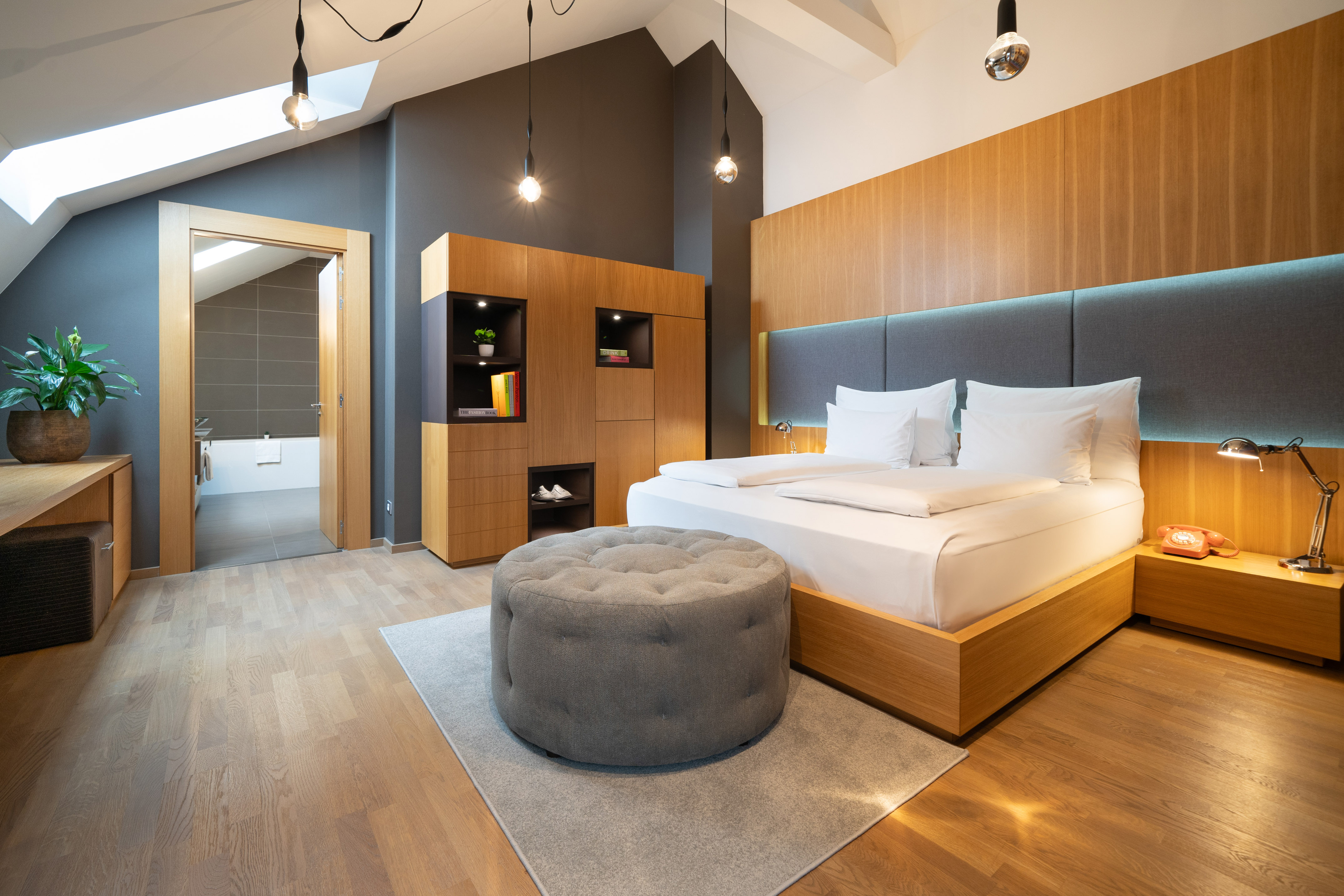 Modern hotel room with wood bed, gray ottoman, skylight, and ensuite bath.