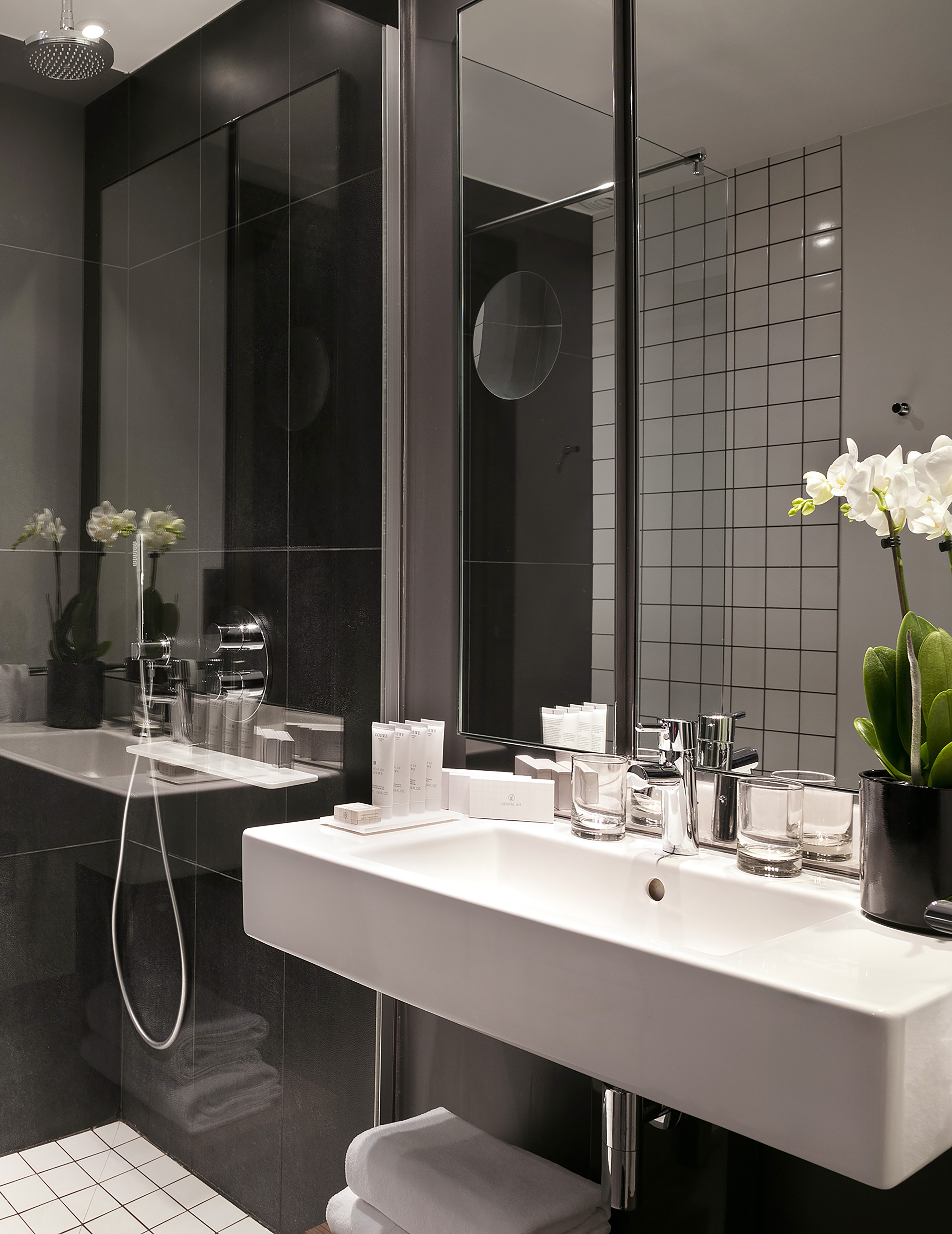Modern hotel bathroom with white sink, chrome faucet, black tiled shower, and orchid.