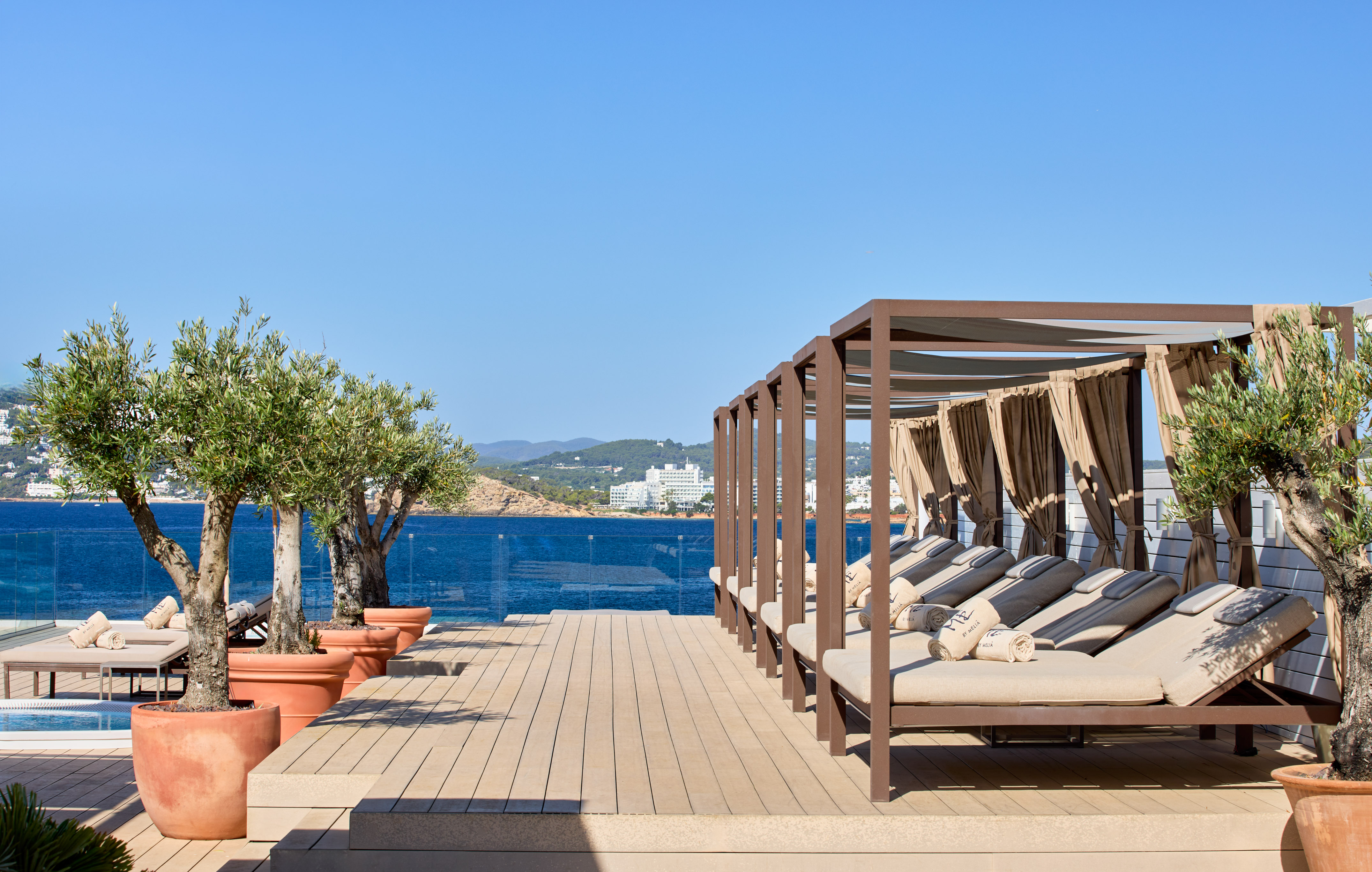 Resort terrace with cabana daybeds, potted olive trees, and a clear sea view.