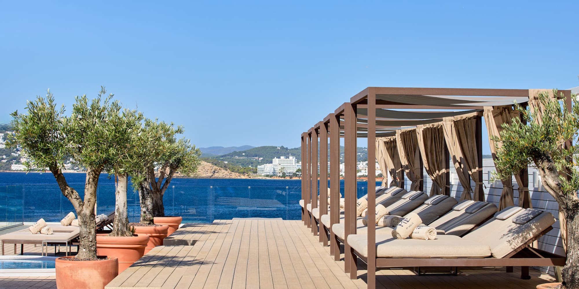 Resort terrace with cabana daybeds, potted olive trees, and a clear sea view.