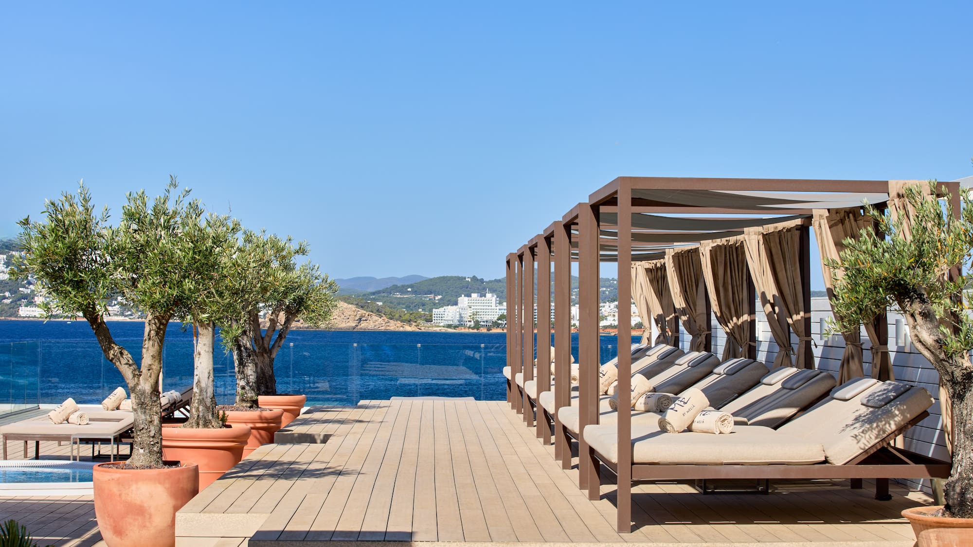 Resort terrace with cabana daybeds, potted olive trees, and a clear sea view.