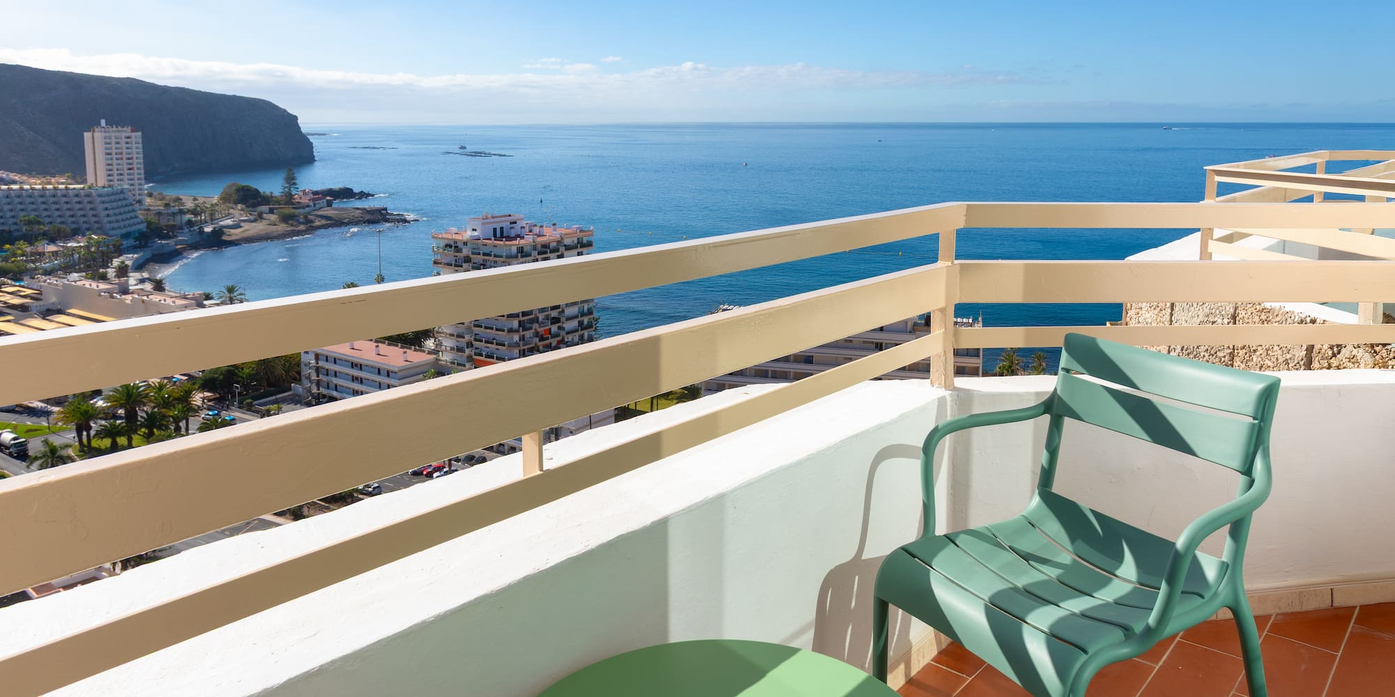 Balcony ocean view: green chair, coastal town, blue sea, cliffs.