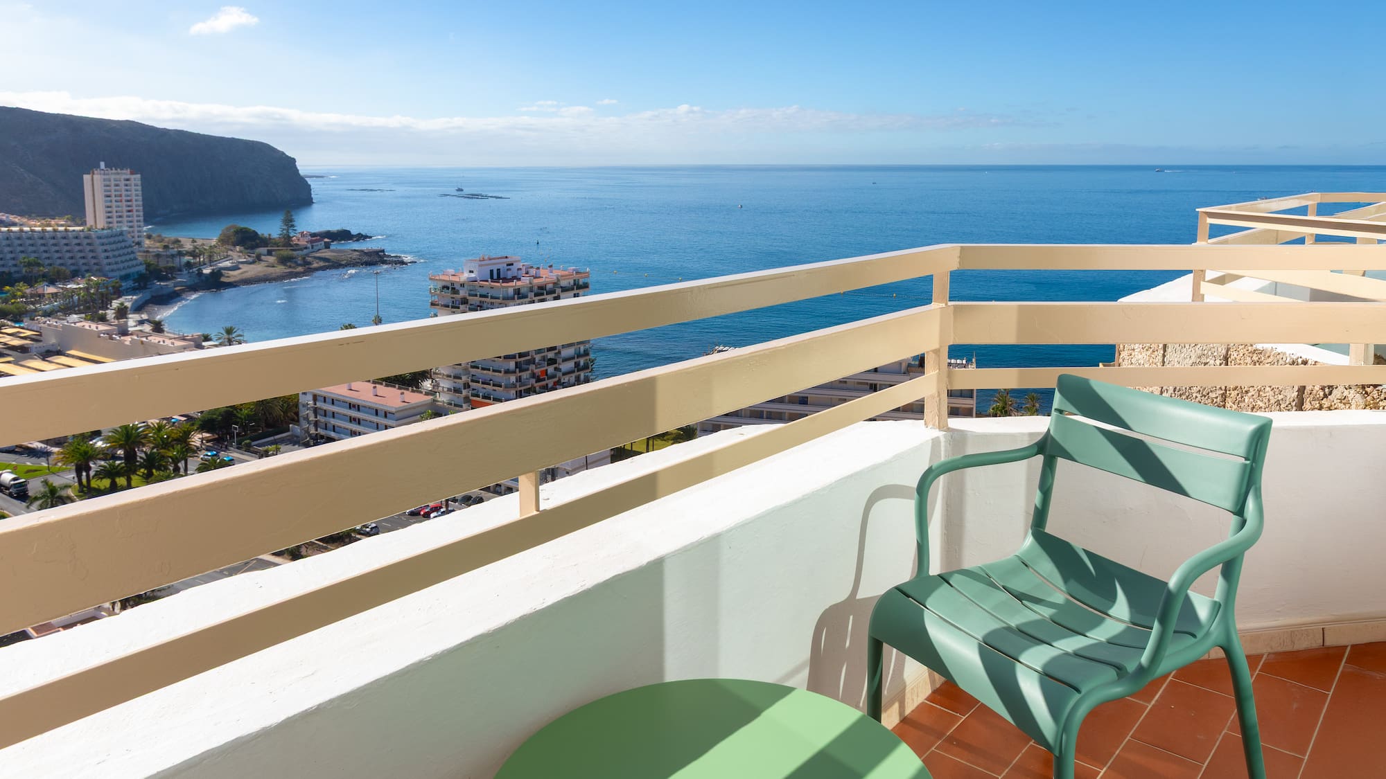 Balcony ocean view: green chair, coastal town, blue sea, cliffs.