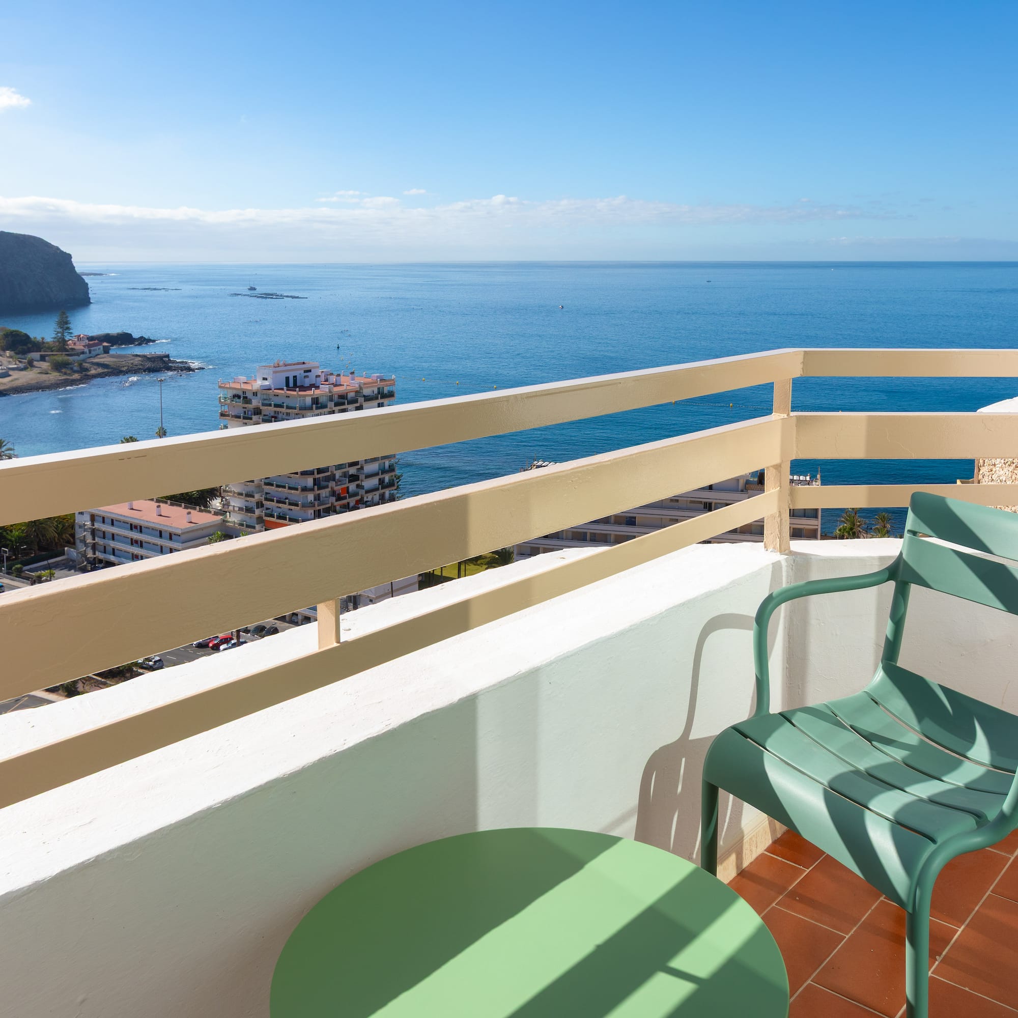 Balcony ocean view: green chair, coastal town, blue sea, cliffs.