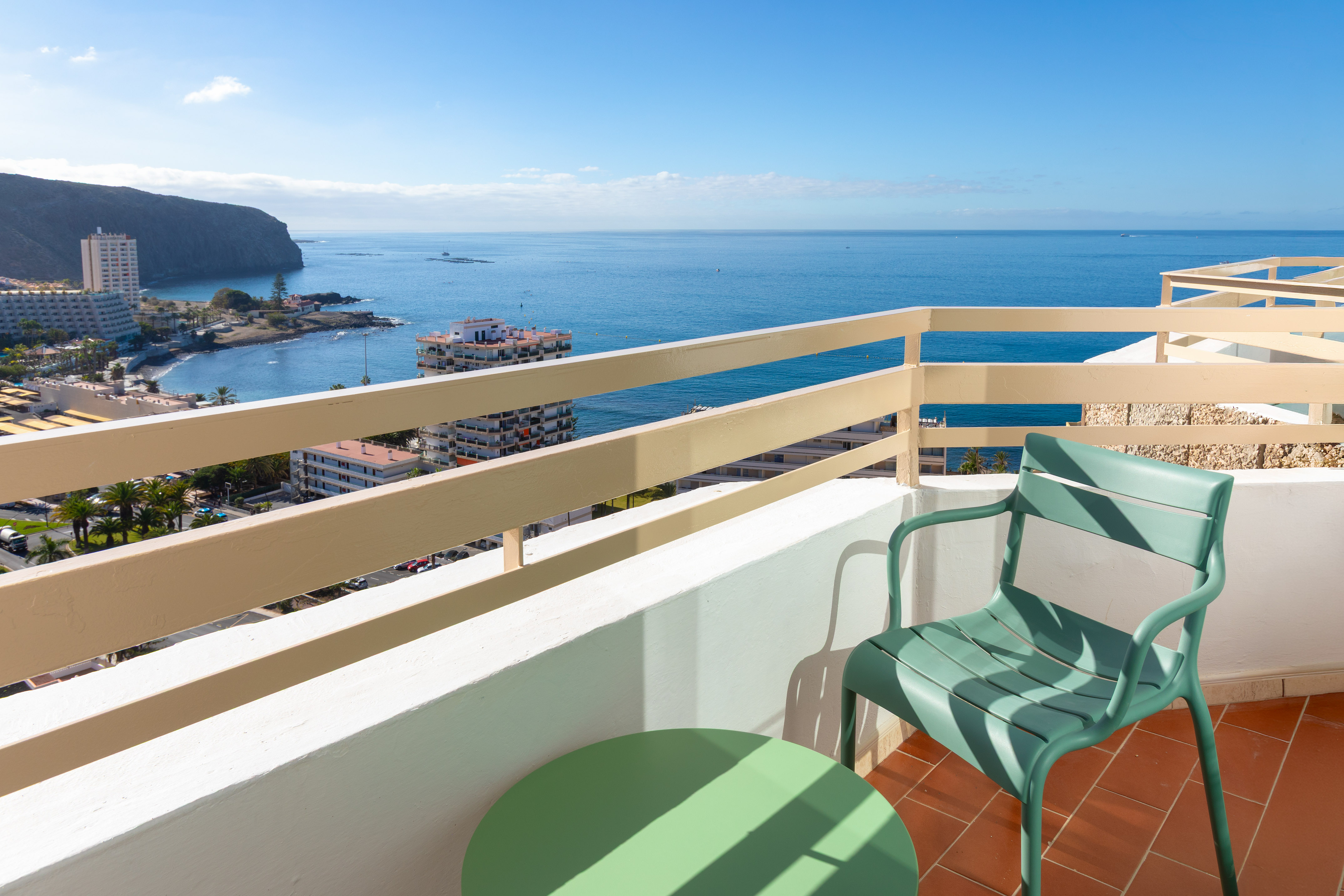 Balcony ocean view: green chair, coastal town, blue sea, cliffs.