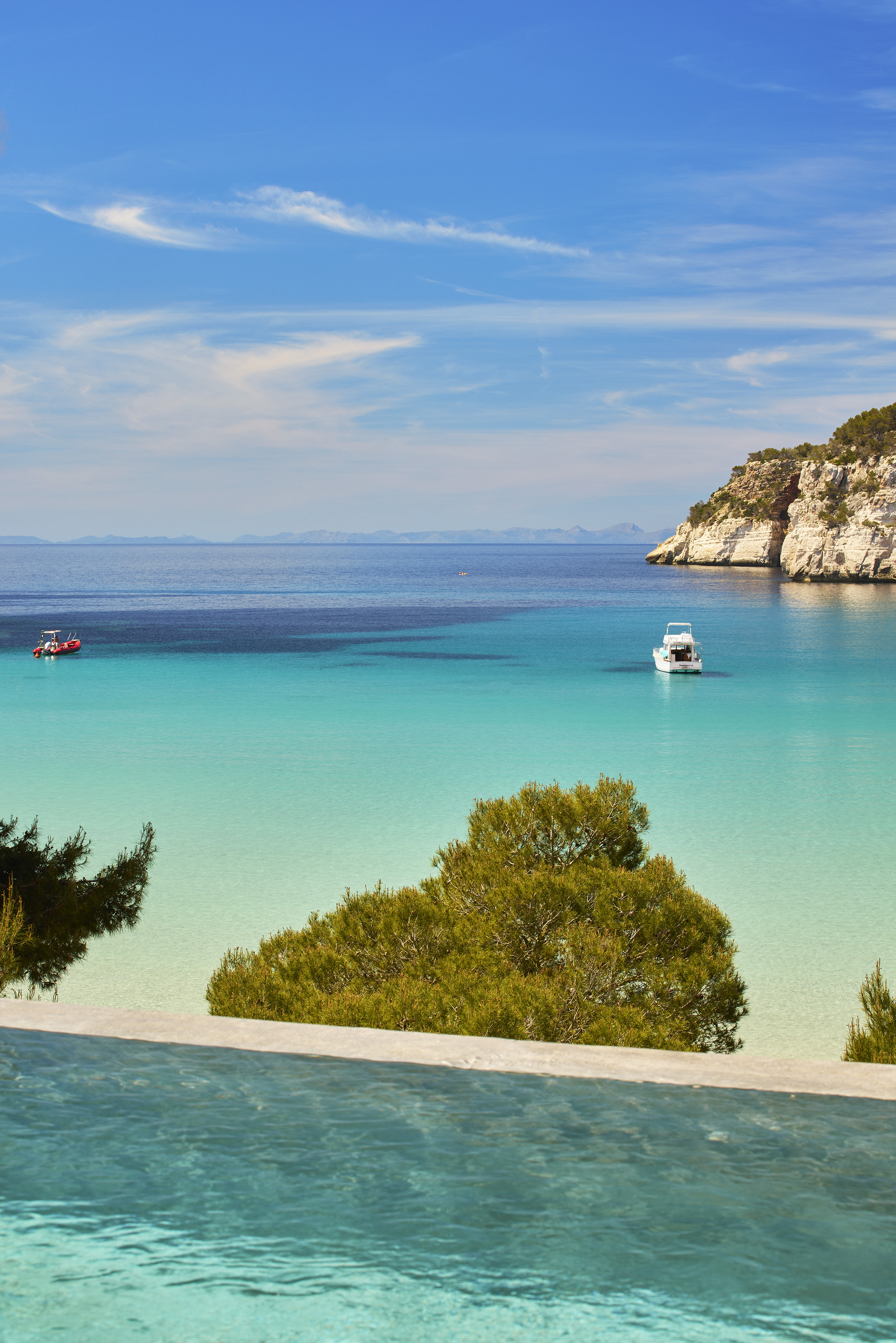 Turquoise Mediterranean bay with boats, rocky cliffs, and infinity pool view.