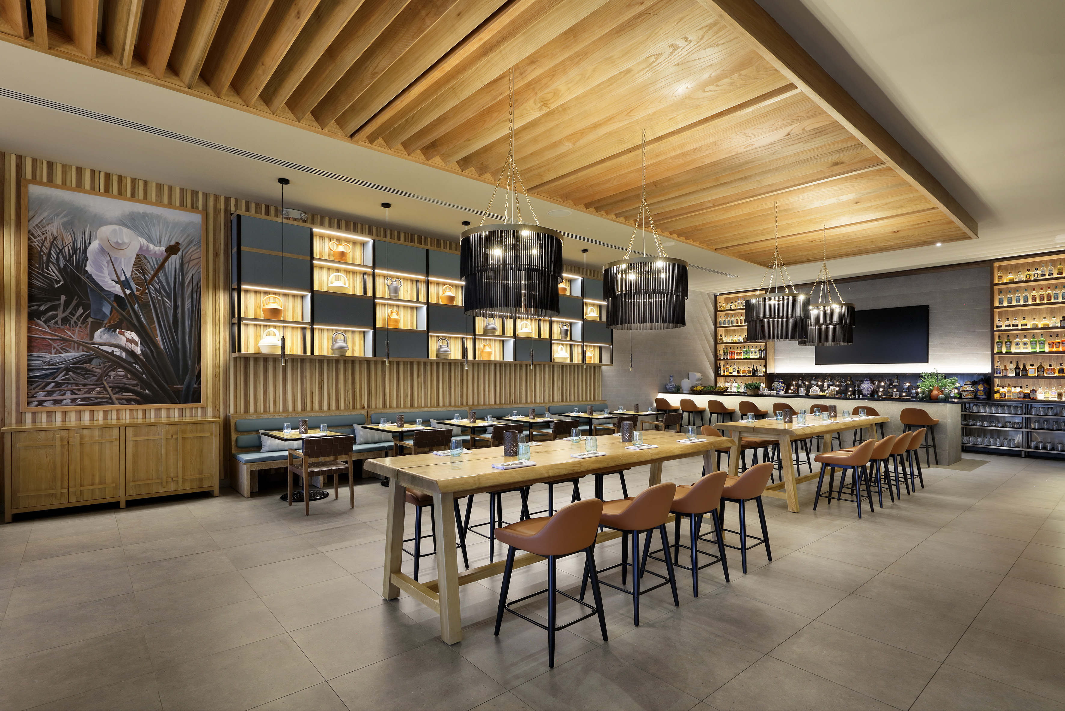 Chic restaurant bar interior with wooden ceiling, communal tables, and agave artwork.