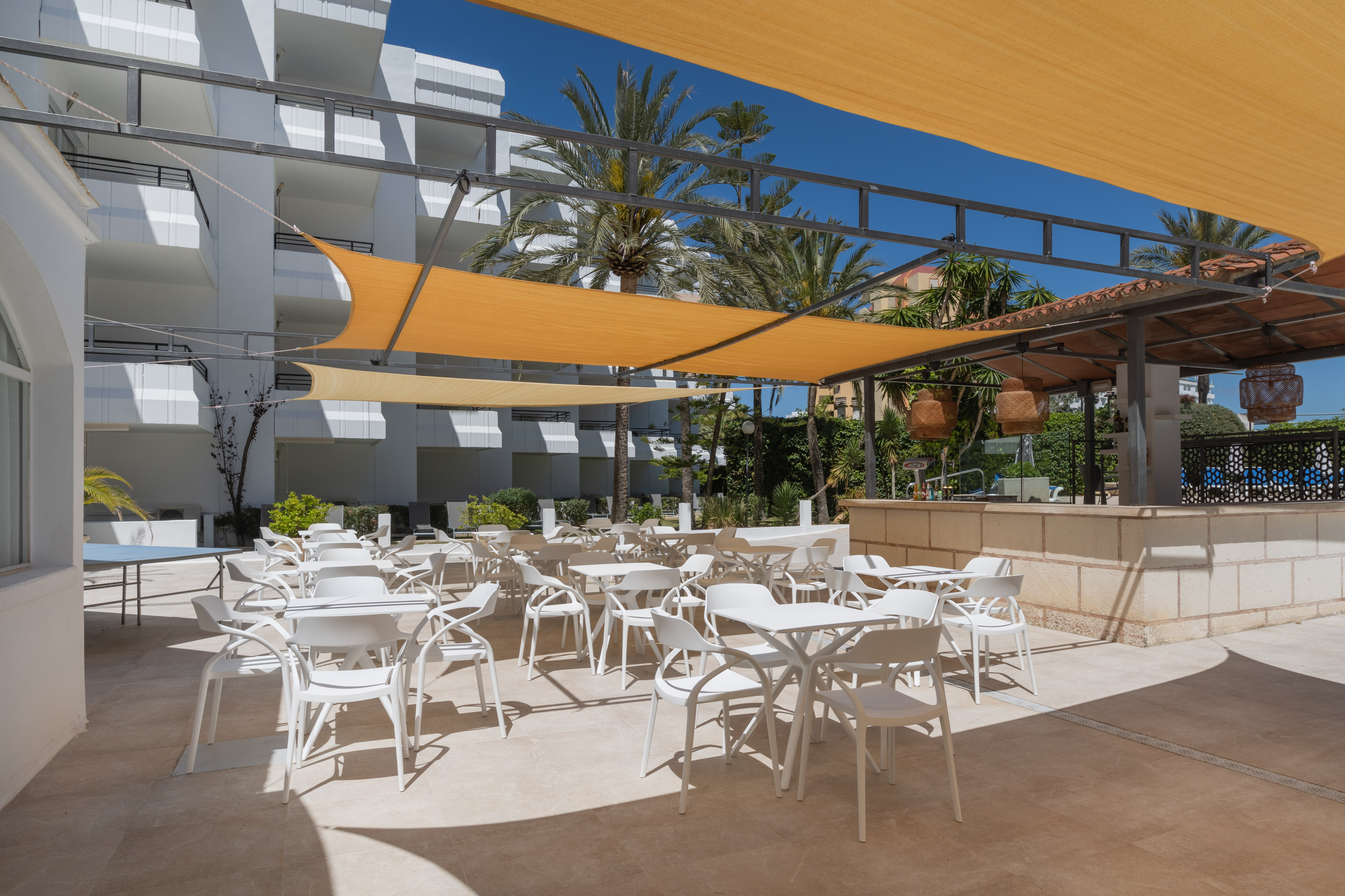 Sunny outdoor hotel terrace with white seating, shade sails, and palm trees.
