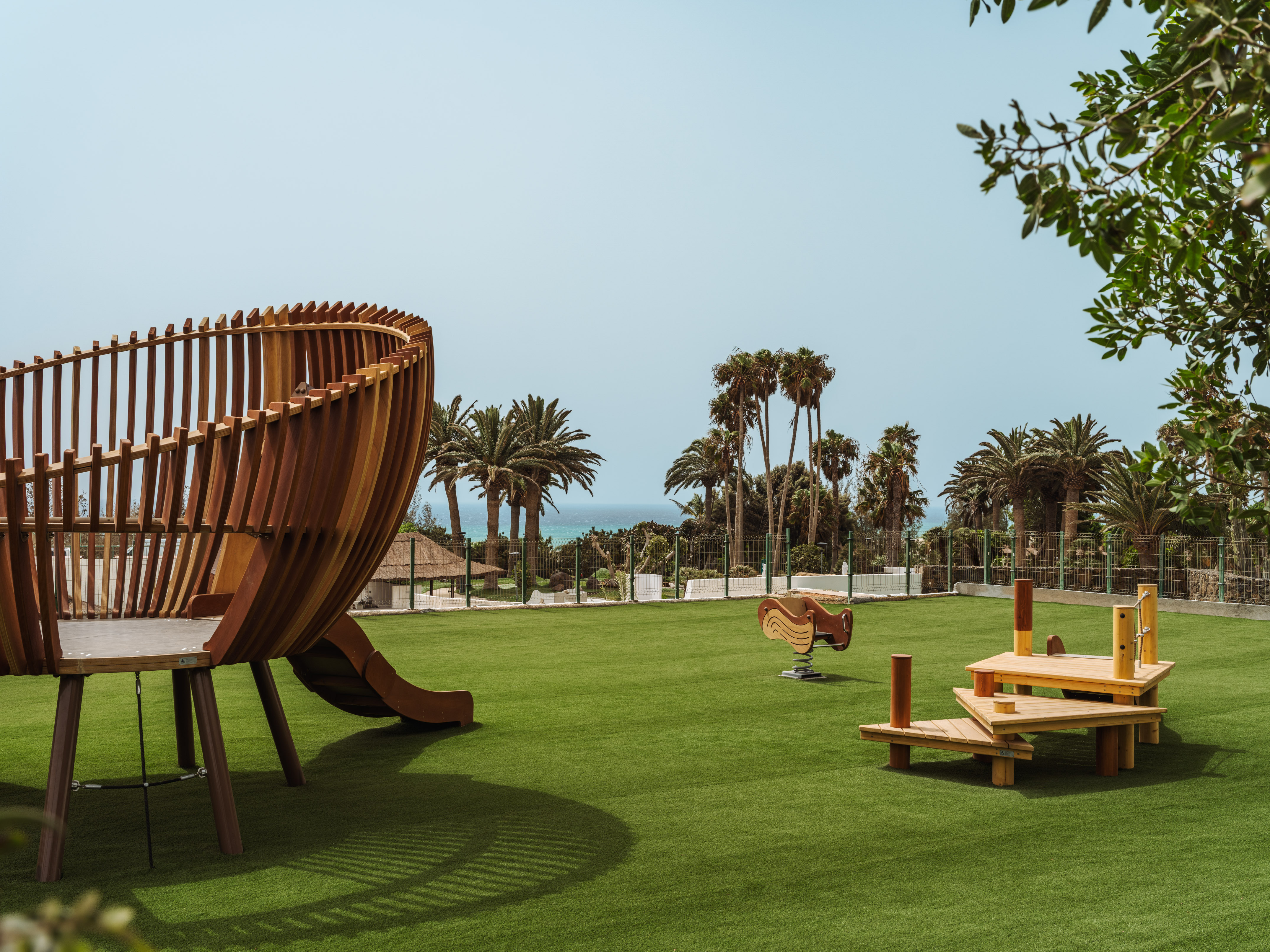 Modern wooden playground on green turf with palm trees and ocean view.