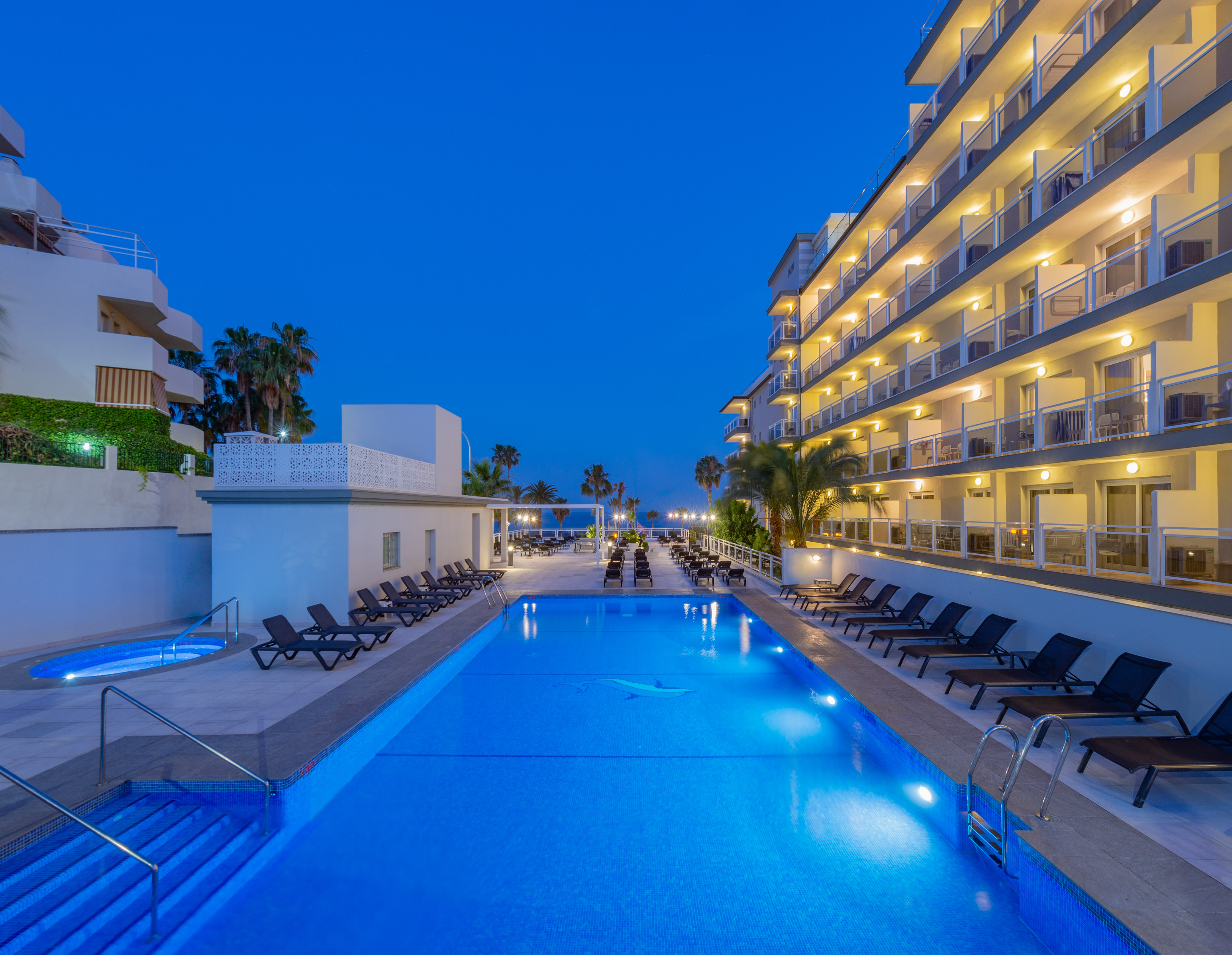 Illuminated resort swimming pool at night with hotel balconies and palm trees.