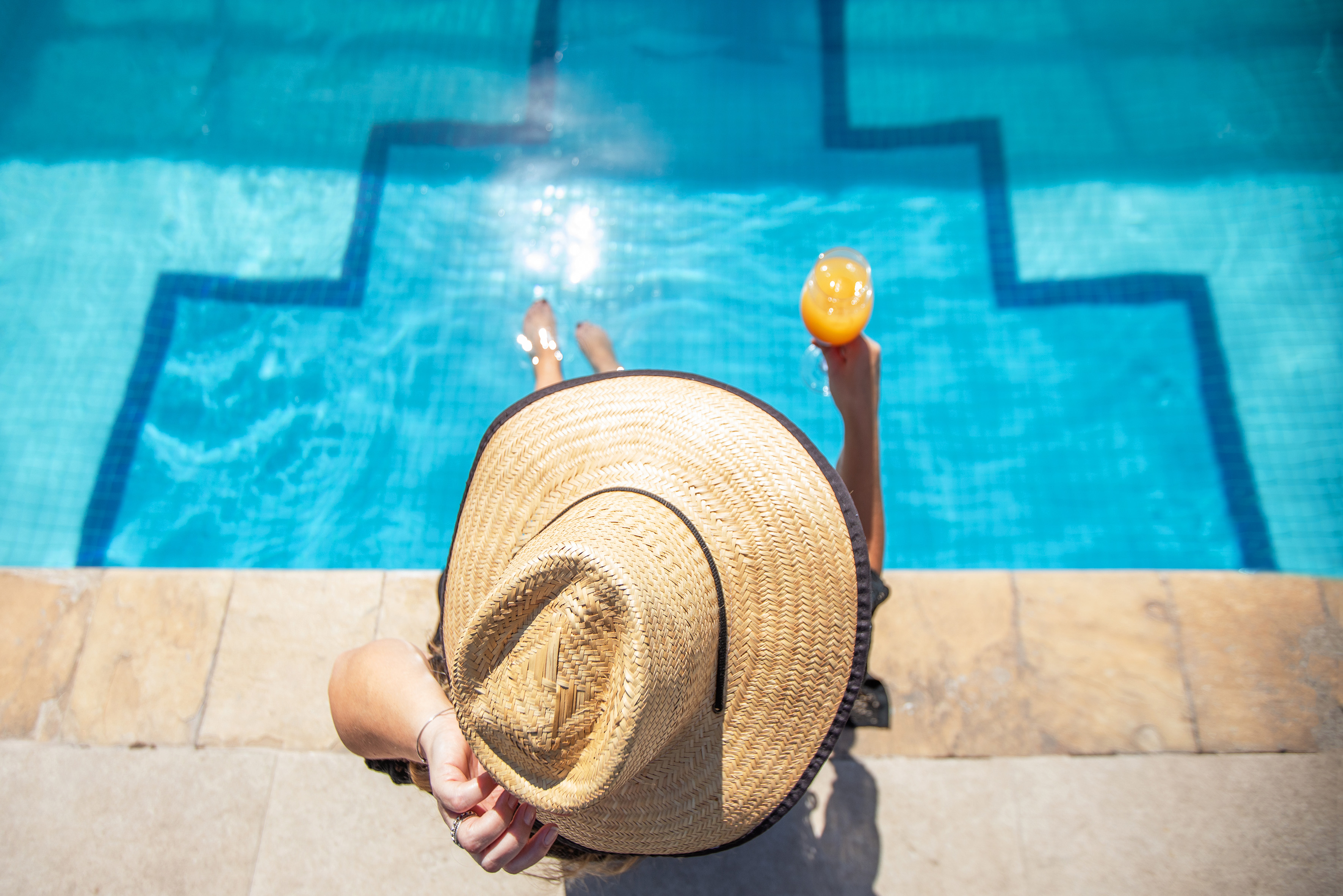 Person in straw hat relaxing poolside, feet in blue pool, holding drink.