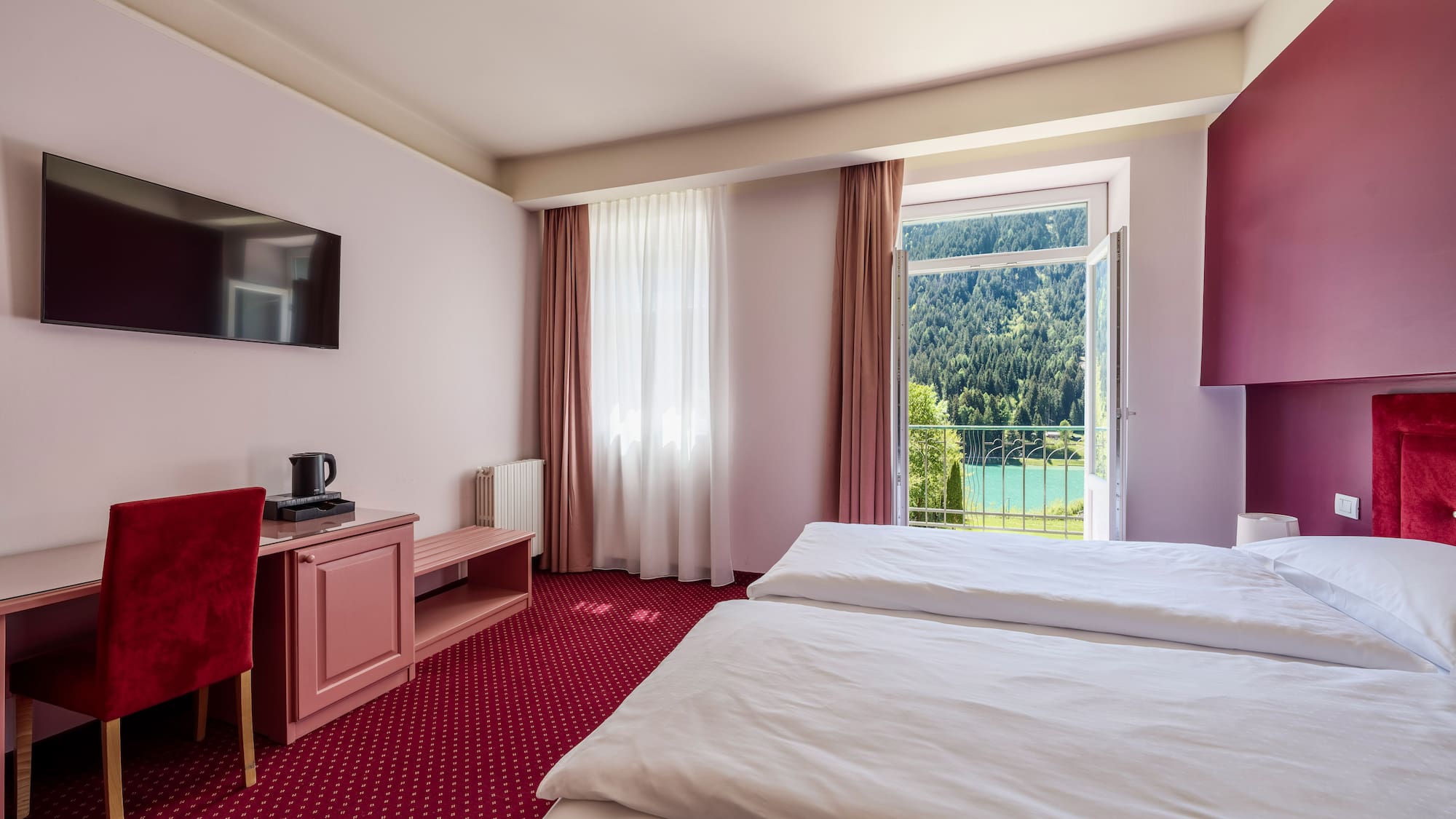 Twin hotel room with desk, TV, and balcony overlooking a mountain lake.