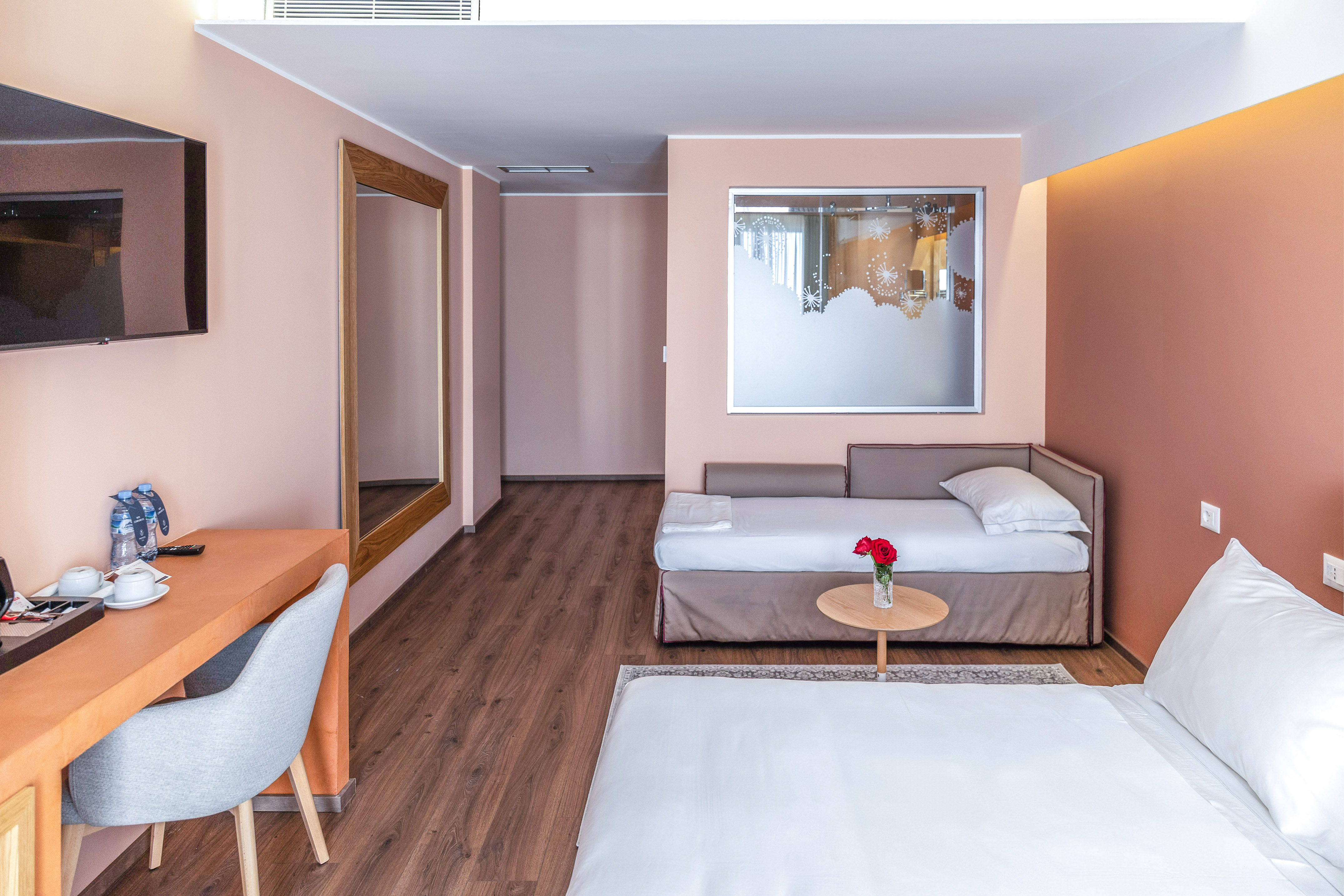 Modern hotel room with bed, sofa bed, work desk, TV, and wooden floor.