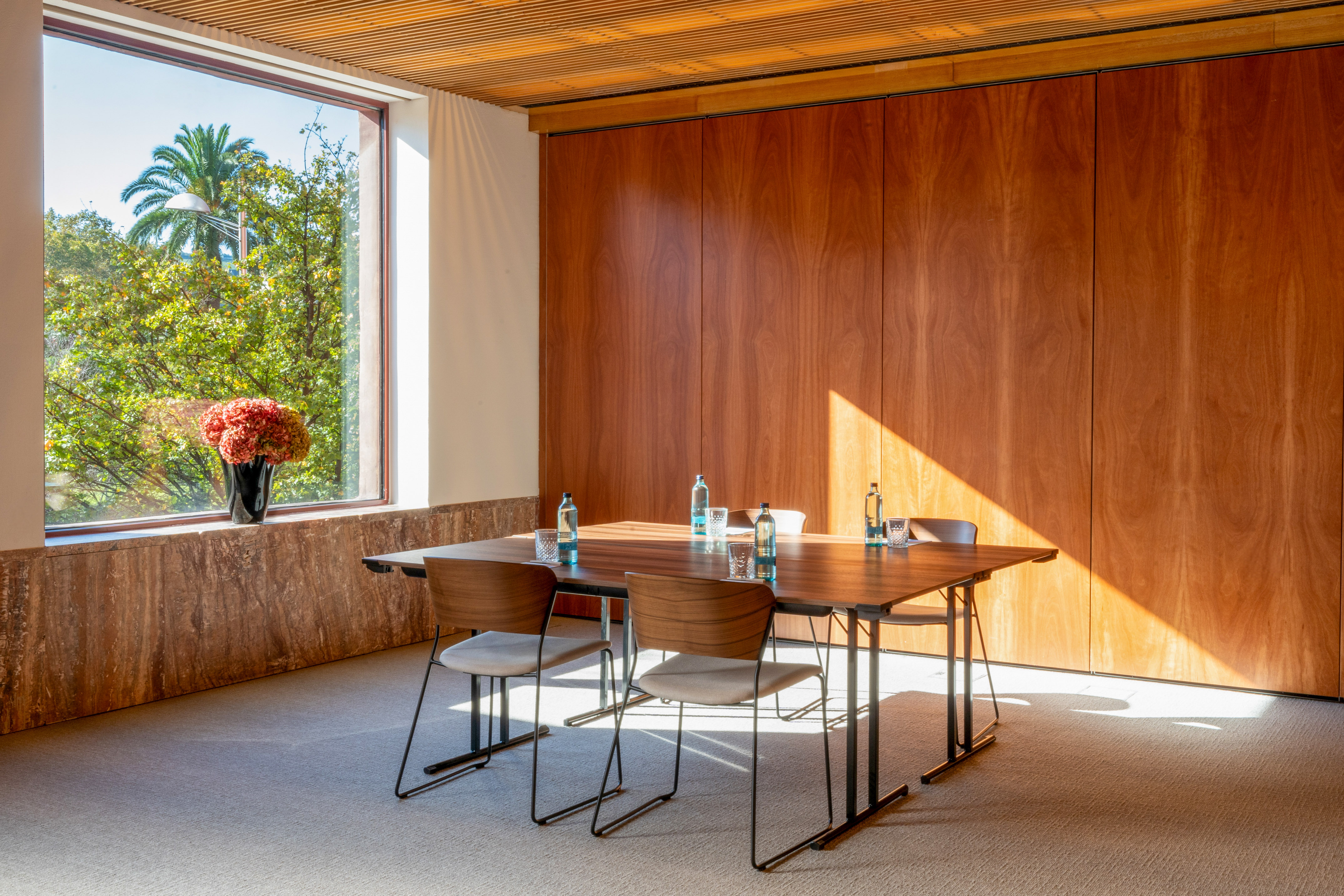 Sunlit modern meeting room with wooden table, chairs, large window, and green trees.