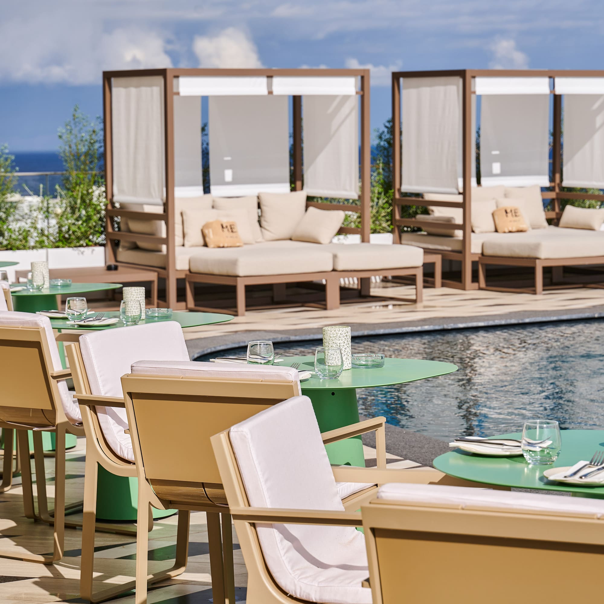 Resort poolside dining area with green tables, beige chairs, daybeds, and ocean view.