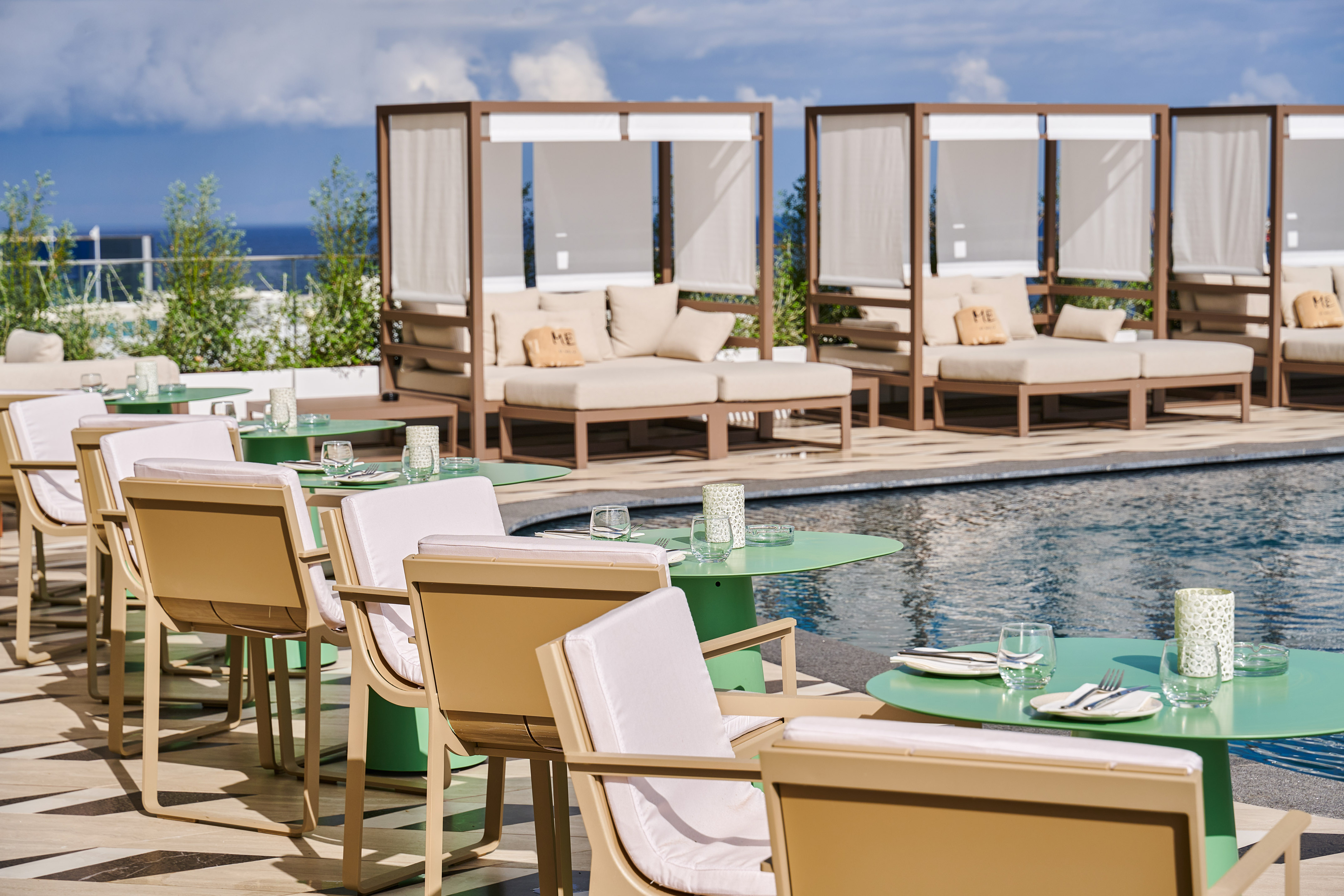 Sunny poolside dining and lounge area with green tables, cabanas, ocean view.
