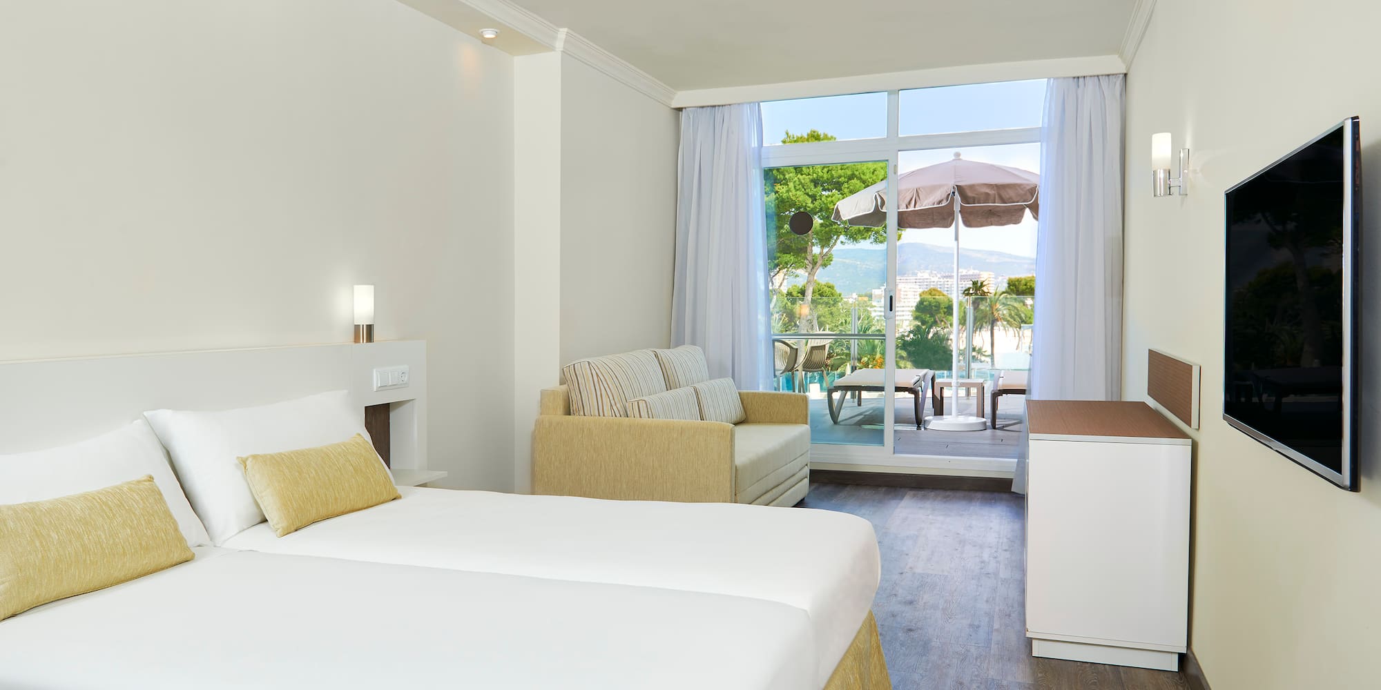 Bright hotel room with beds, sofa, TV, and balcony with scenic sea and mountain views.