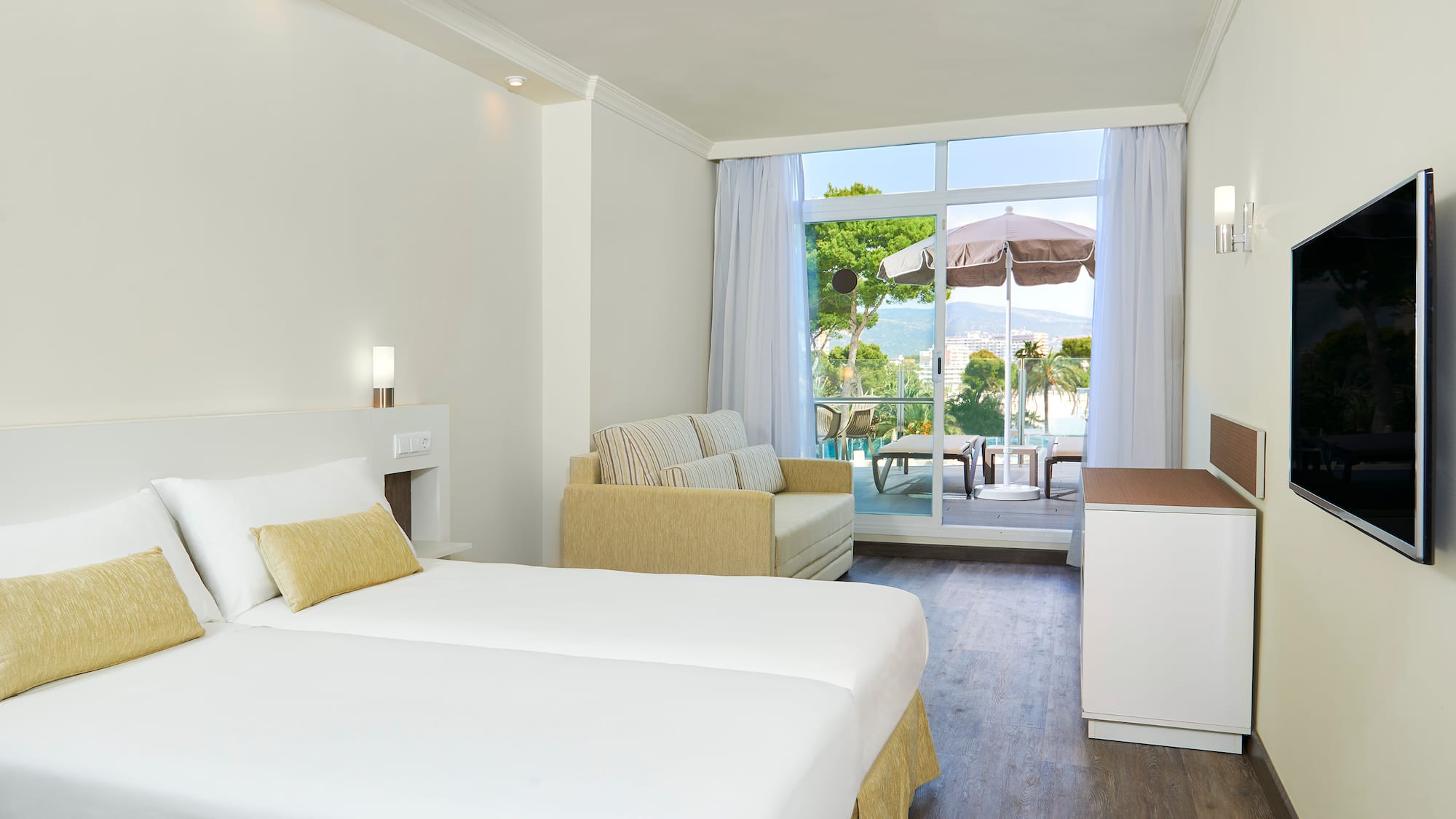 Bright hotel room with beds, sofa, TV, and balcony with scenic sea and mountain views.