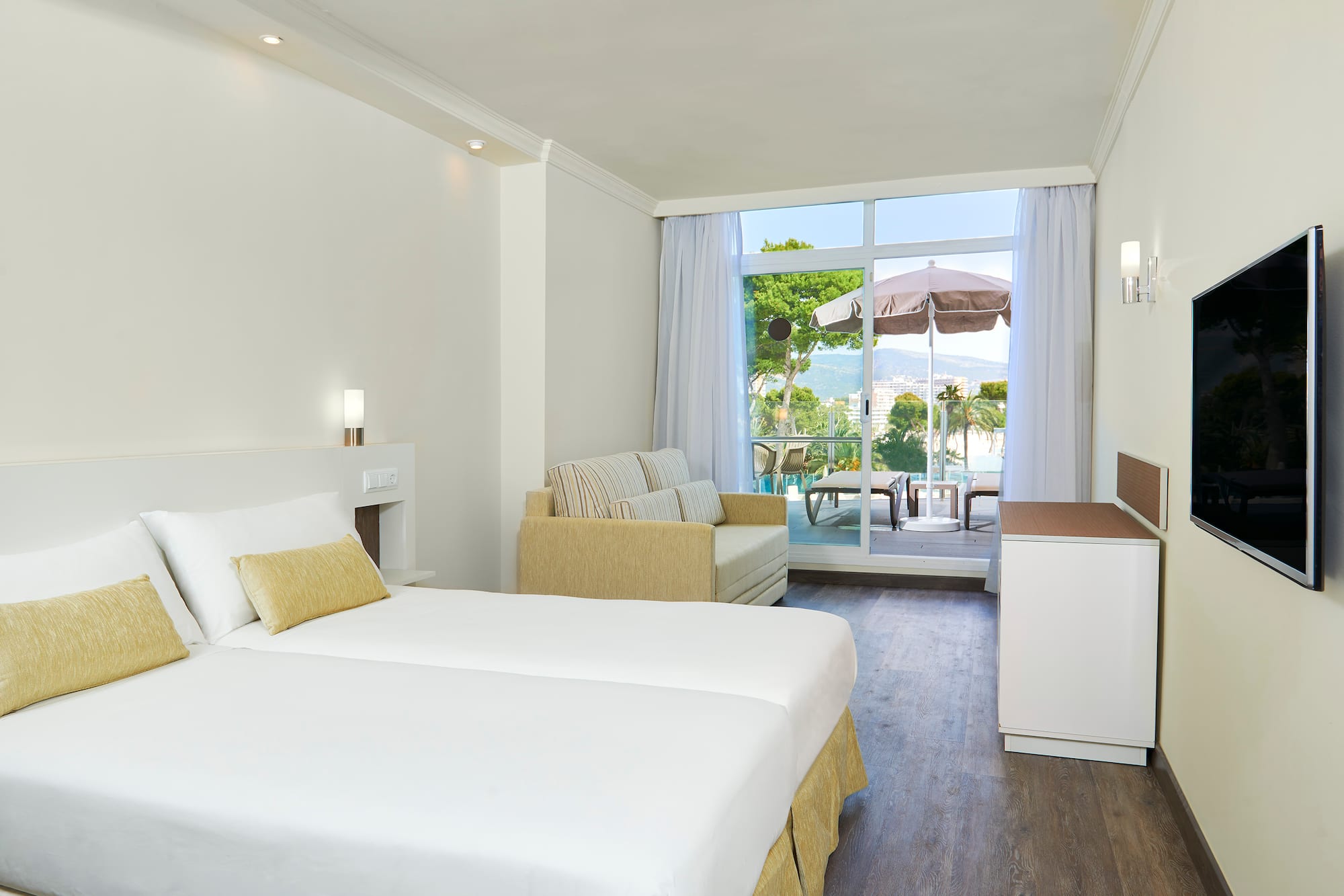 Bright hotel room with beds, sofa, TV, and balcony with scenic sea and mountain views.