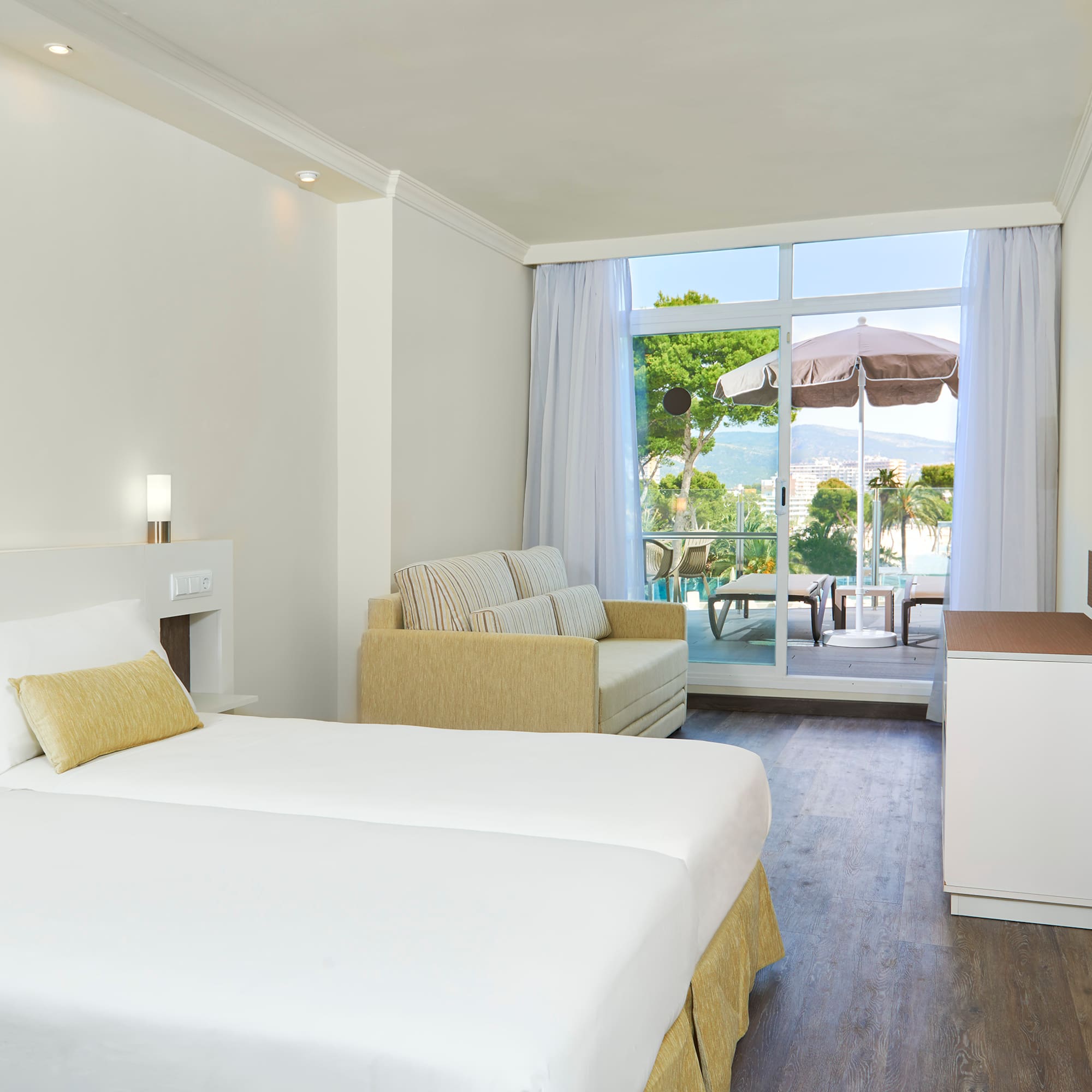 Bright hotel room with beds, sofa, TV, and balcony with scenic sea and mountain views.