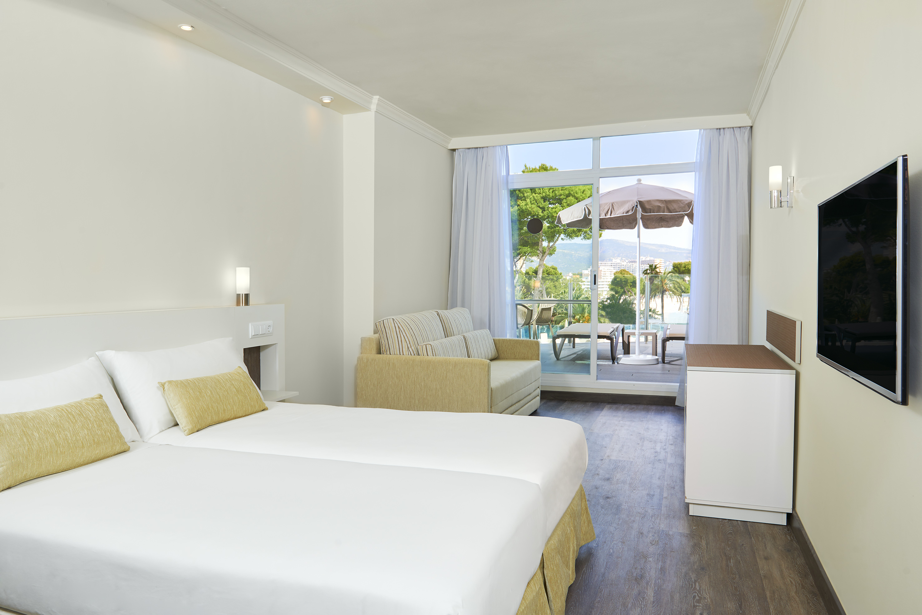 Bright hotel room with beds, sofa, TV, and balcony with scenic sea and mountain views.