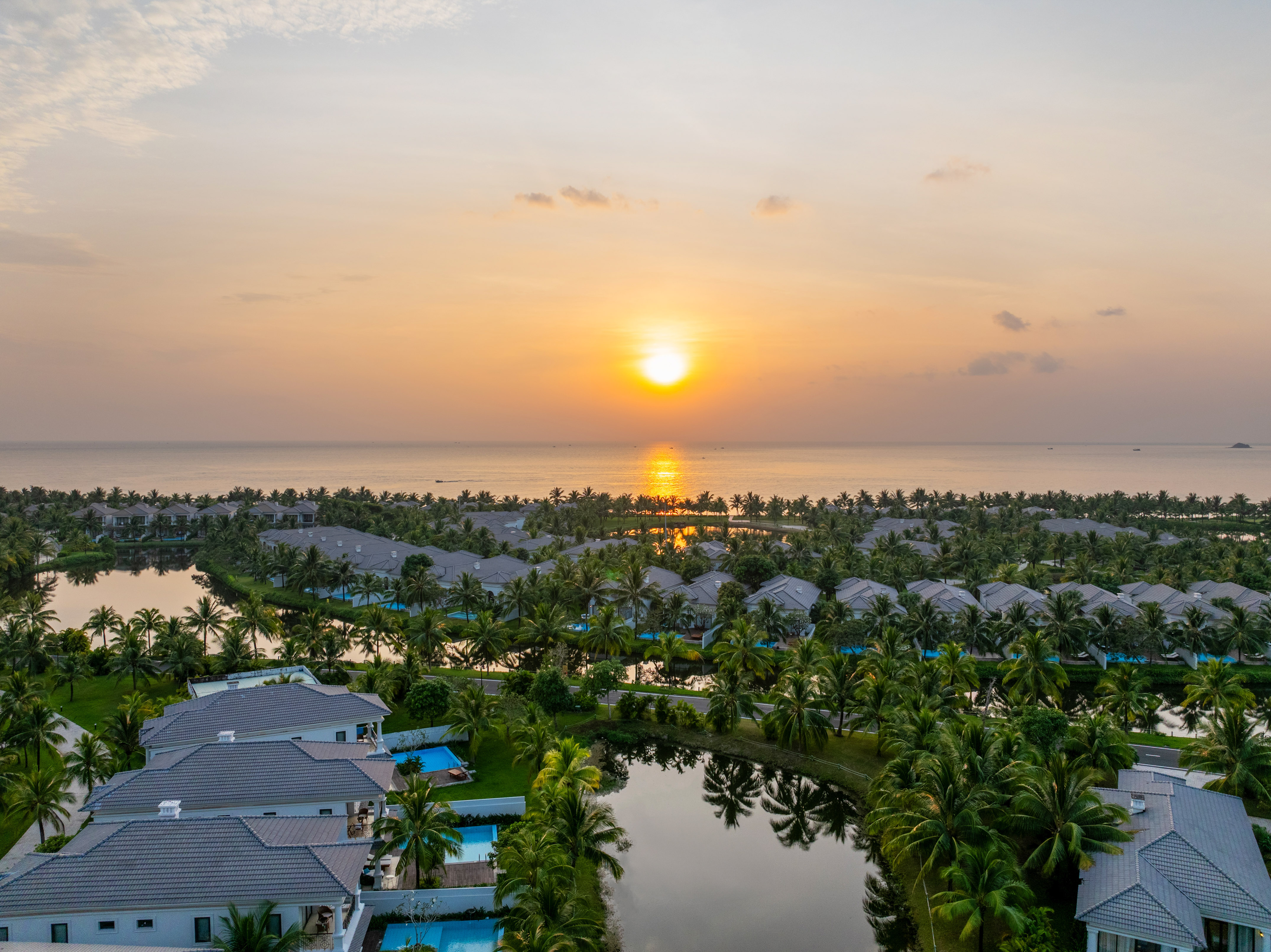 Tropical resort villas with palm trees and lakes, ocean sunset aerial view.