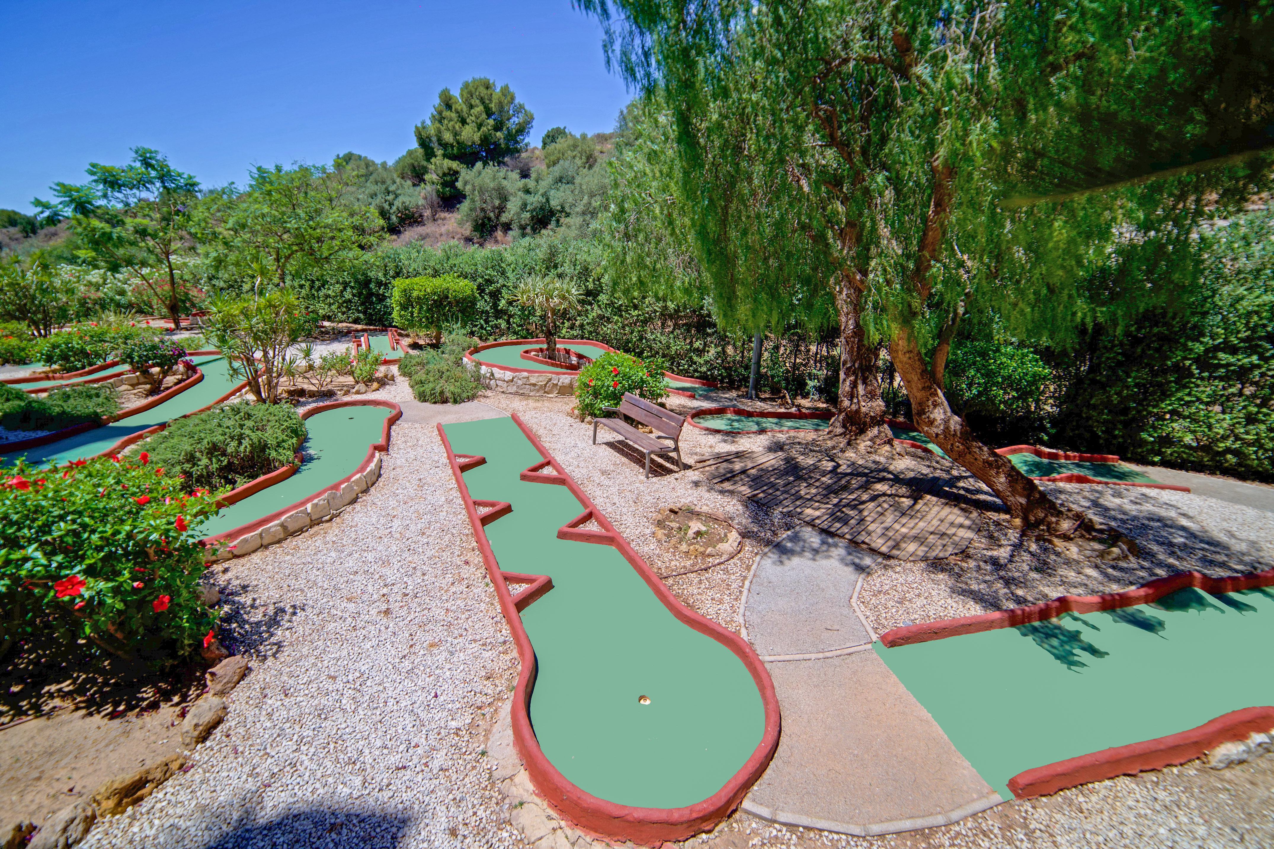 Sunny outdoor mini-golf course with green tracks, gravel, and surrounding trees.
