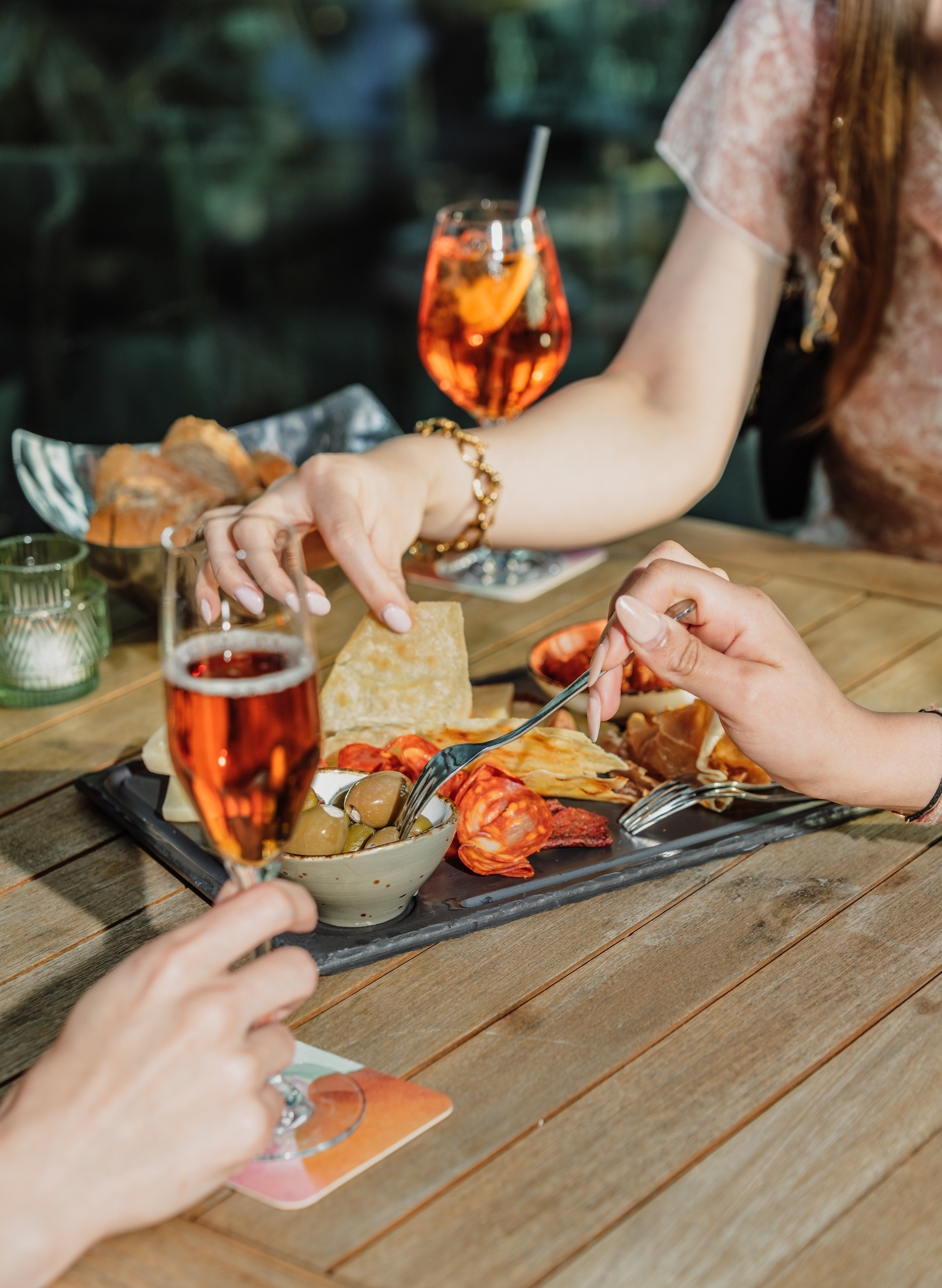 Hands sharing charcuterie board with olives, cured meats, cheese, and cocktails.