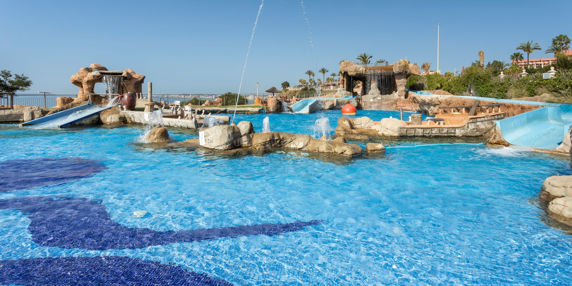 Water park pool with slides, rock waterfalls, fountains, and floor mosaics.