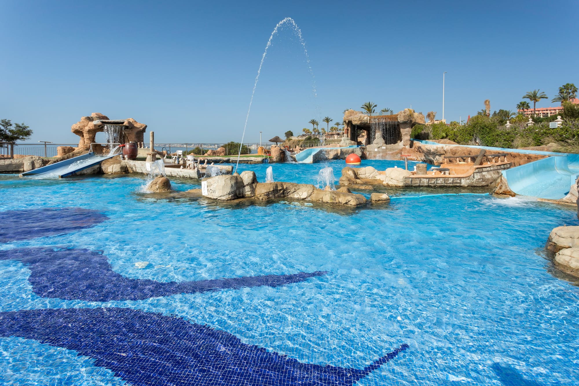 Water park pool with slides, rock waterfalls, fountains, and floor mosaics.
