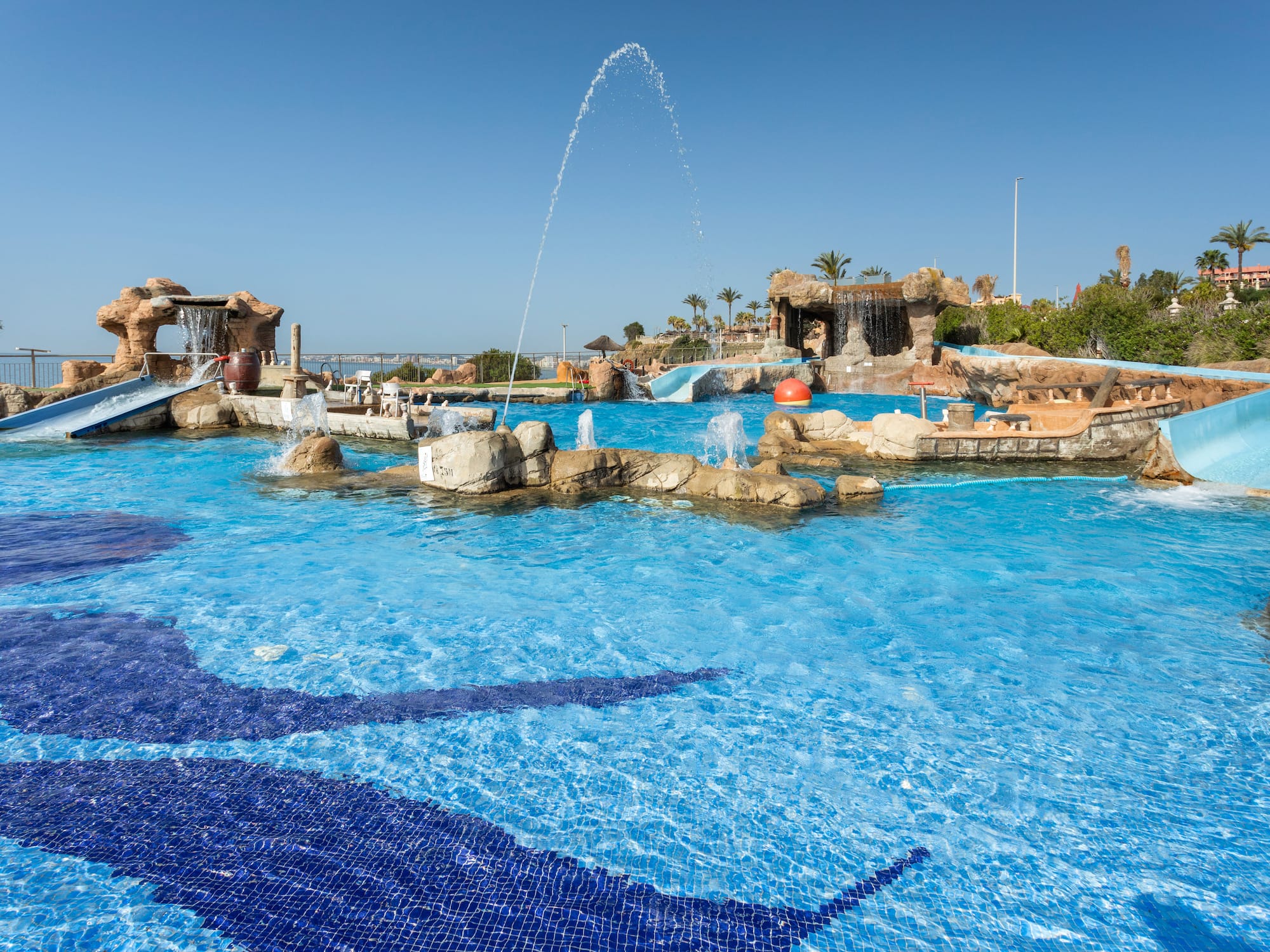 Sunny water park pool with slides, fountains, waterfalls, and blue mosaic tiles.