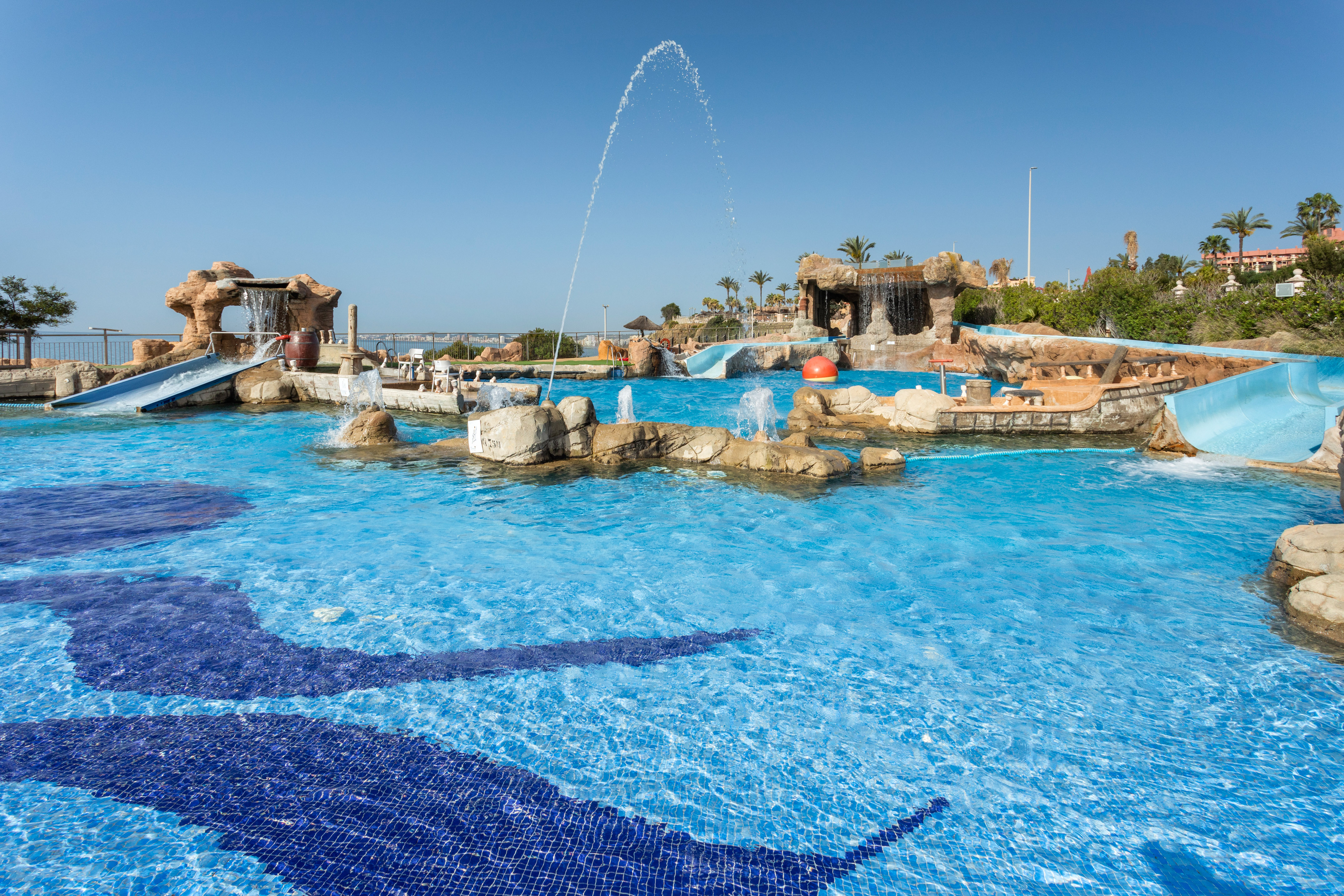 Water park pool with slides, rock waterfalls, fountains, and floor mosaics.