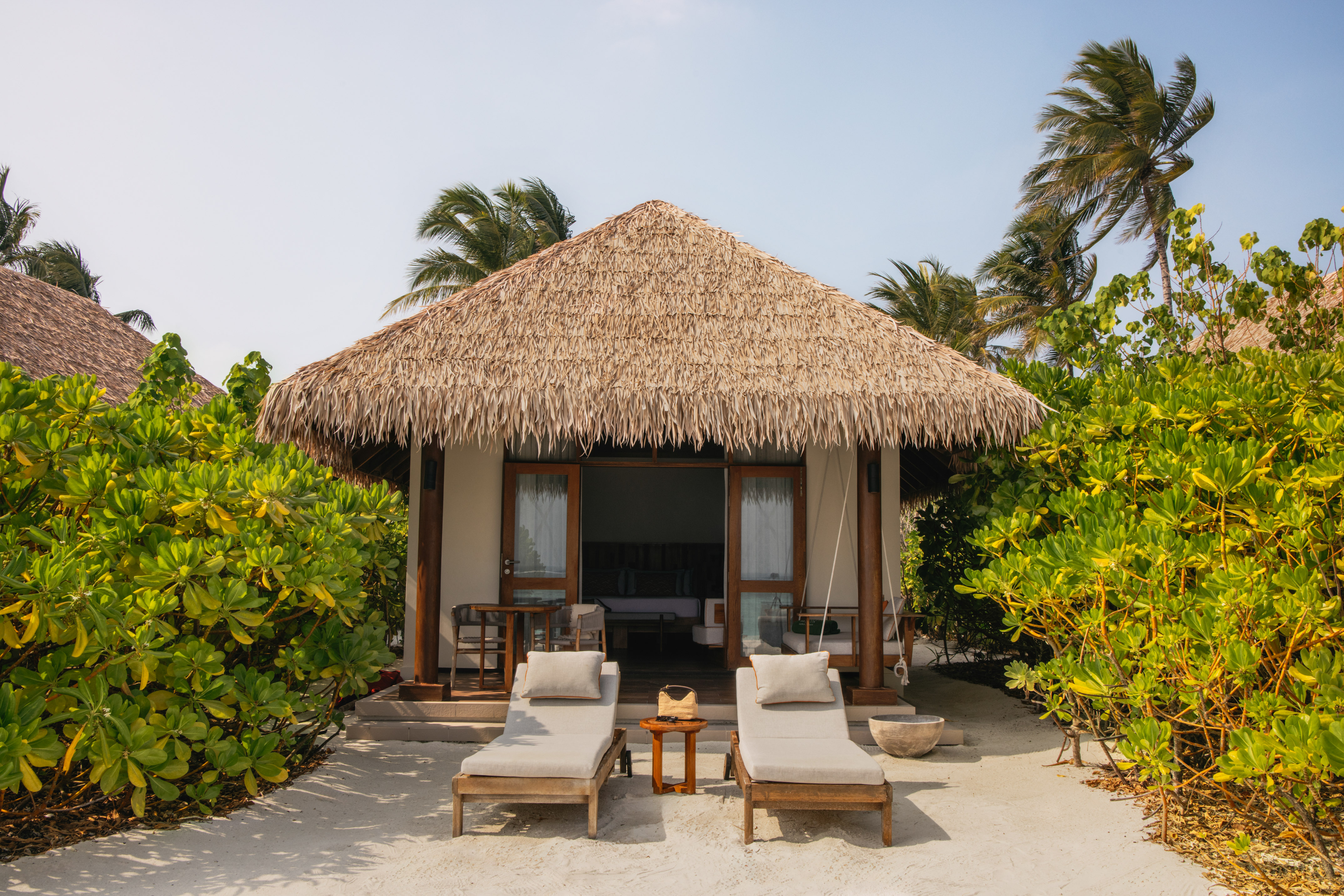 Maldives beach bungalow with thatched roof, sun loungers, and palm trees.