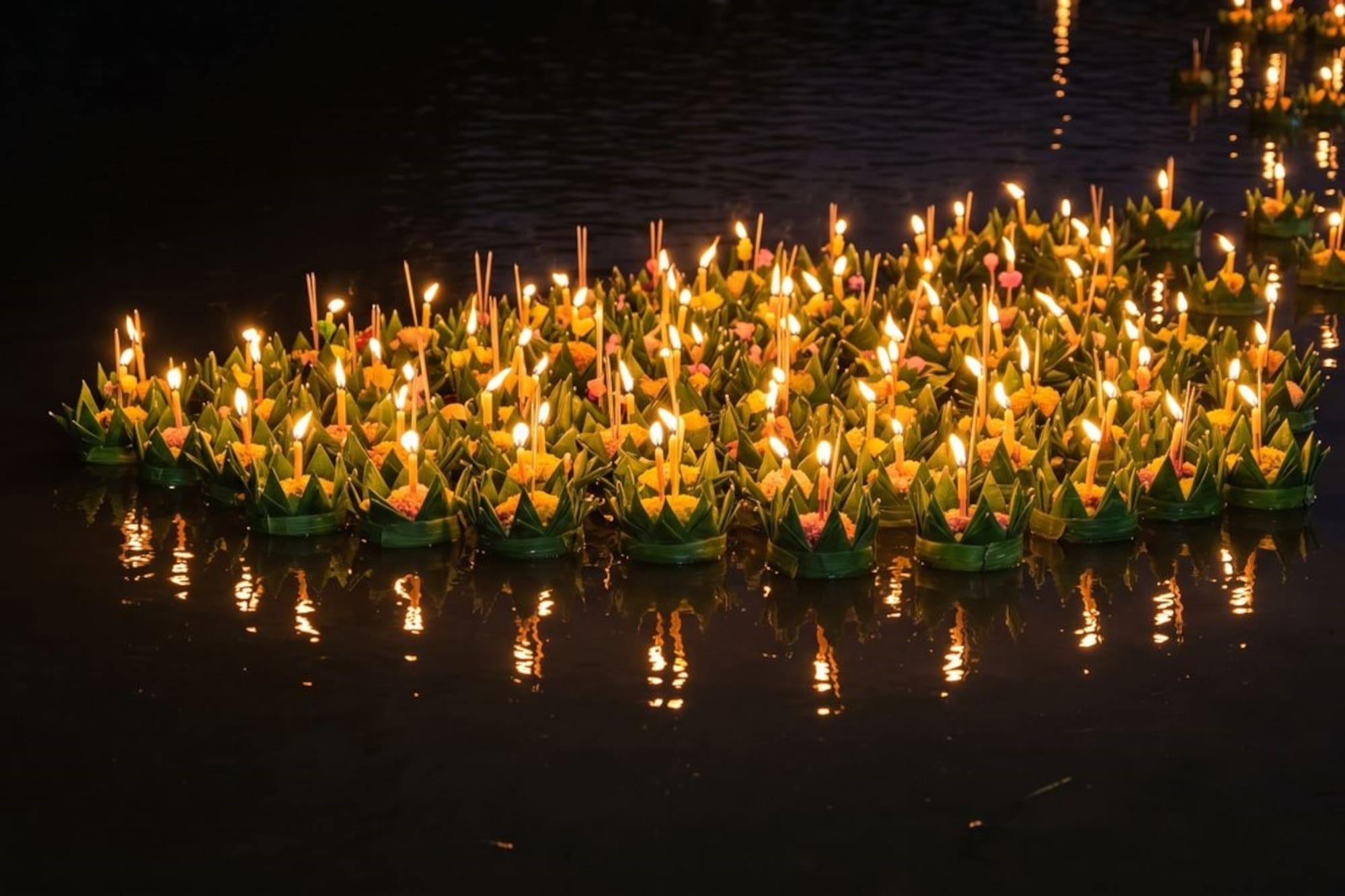 Floating krathongs with lit candles and yellow flowers on dark water.