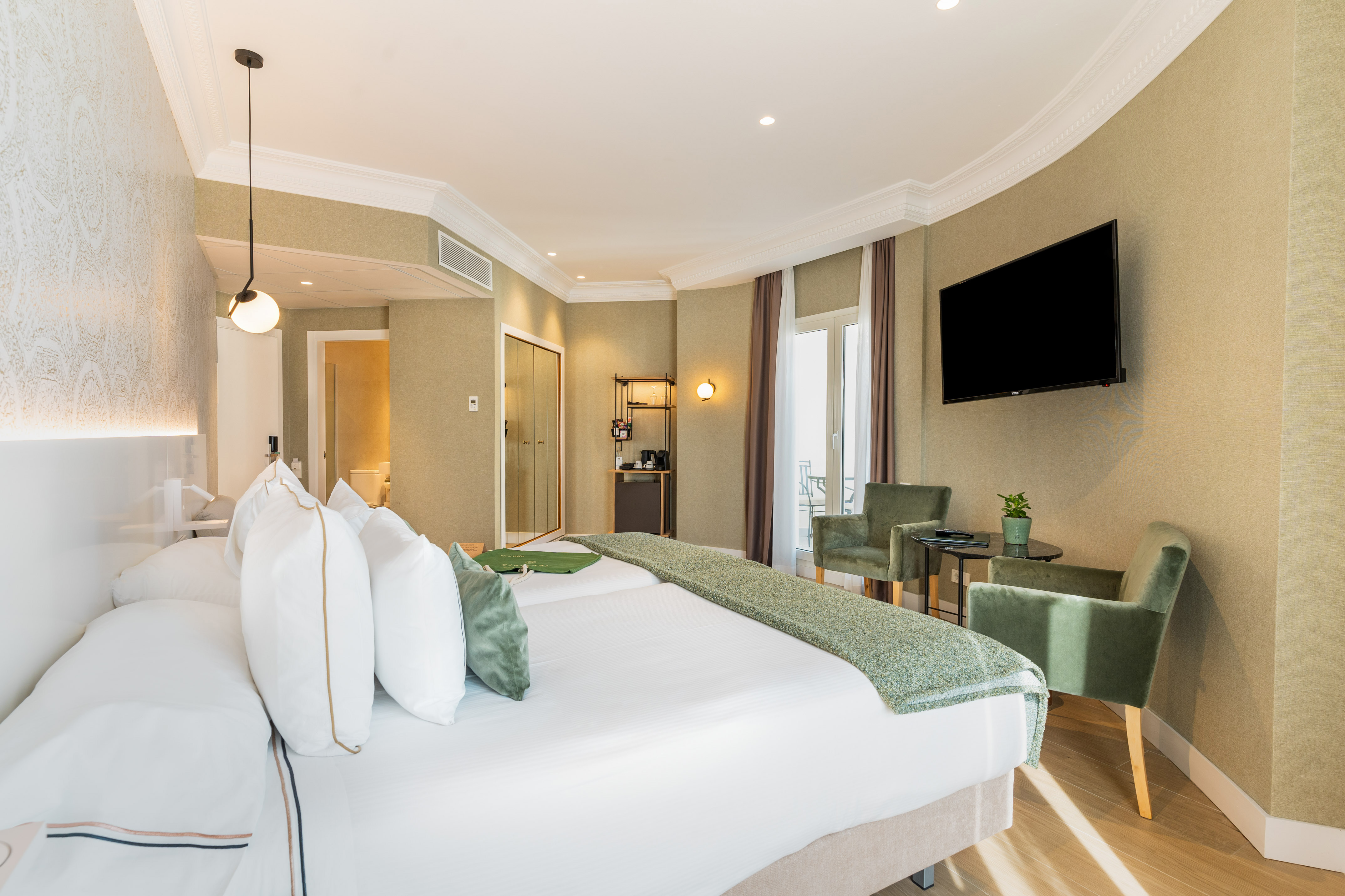 Modern hotel bedroom with large bed, green chairs, TV, and balcony.