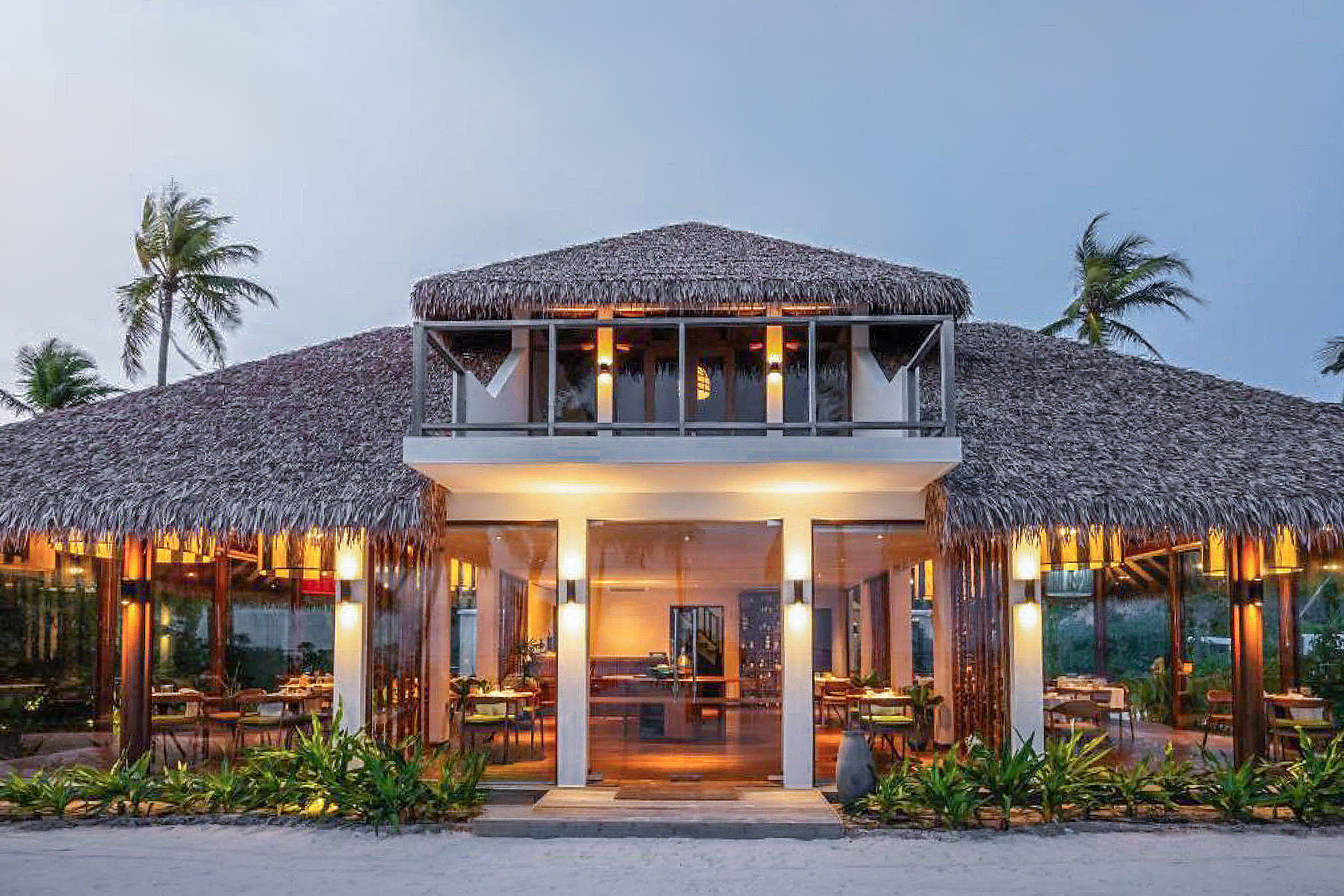 Tropical resort restaurant with thatched roof, glass walls, and evening lighting.