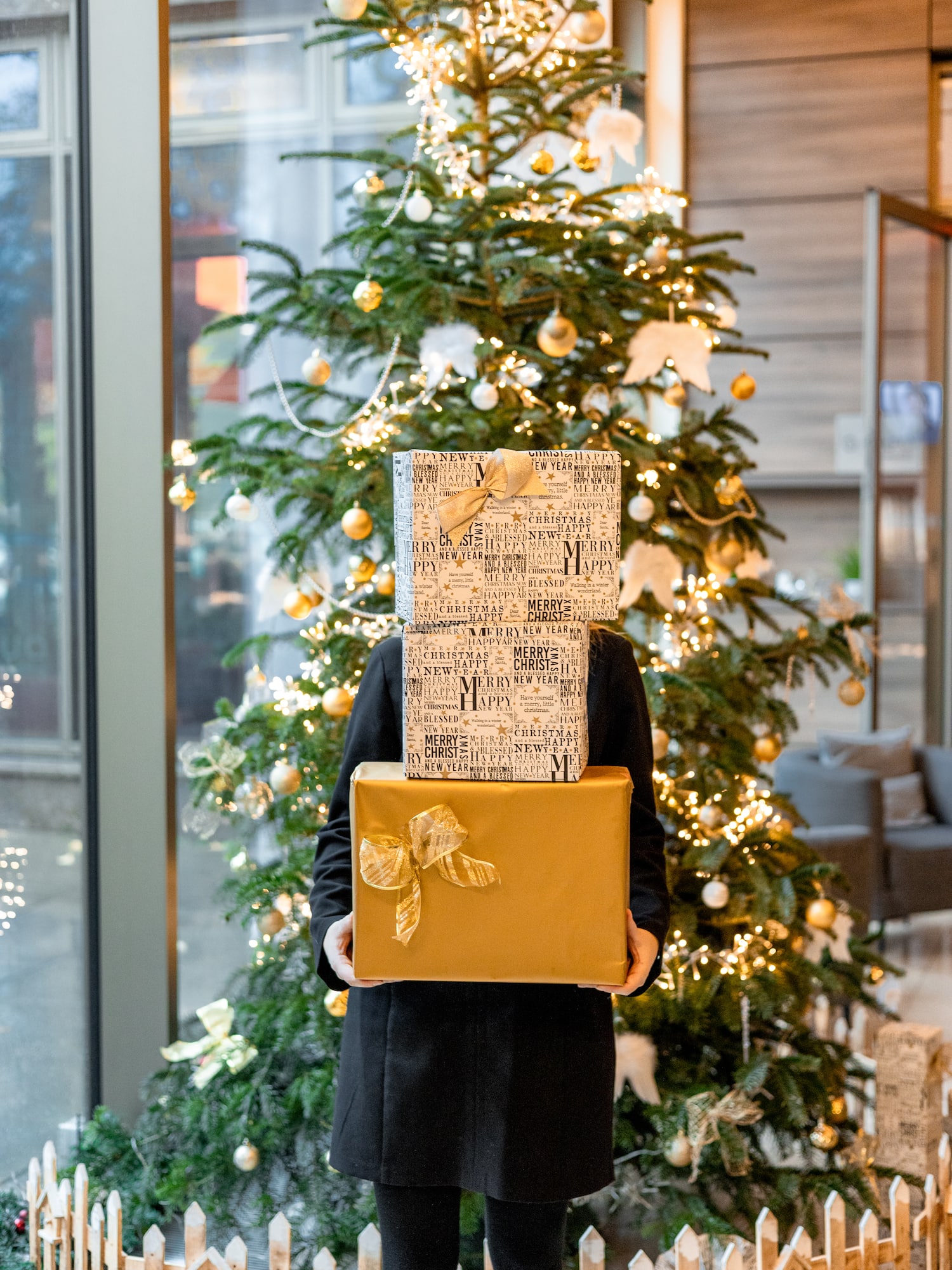 Person holding festive stacked Christmas presents near a lit Christmas tree.
