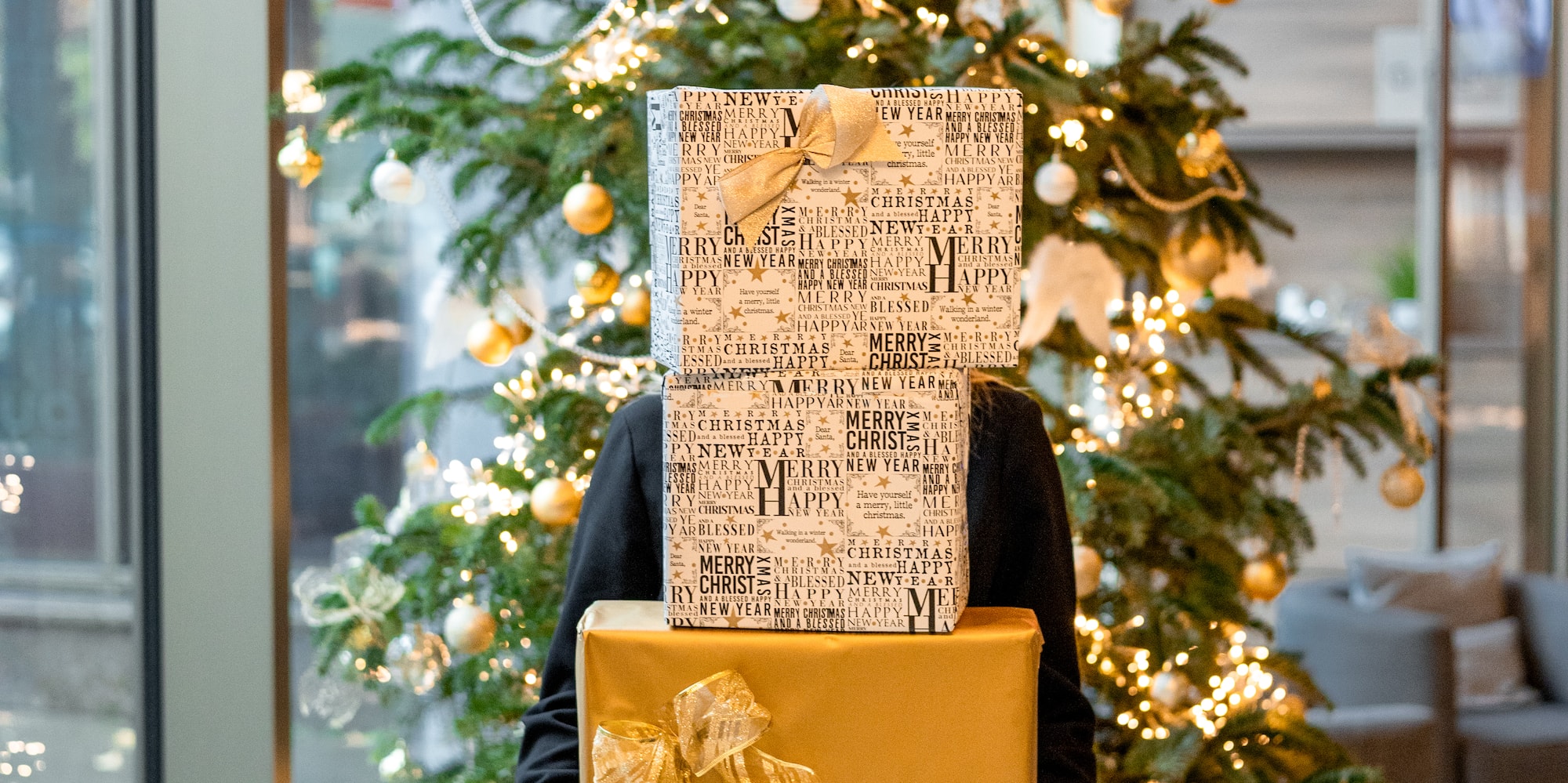 Person holds gold & festive presents in front of a glowing Christmas tree.