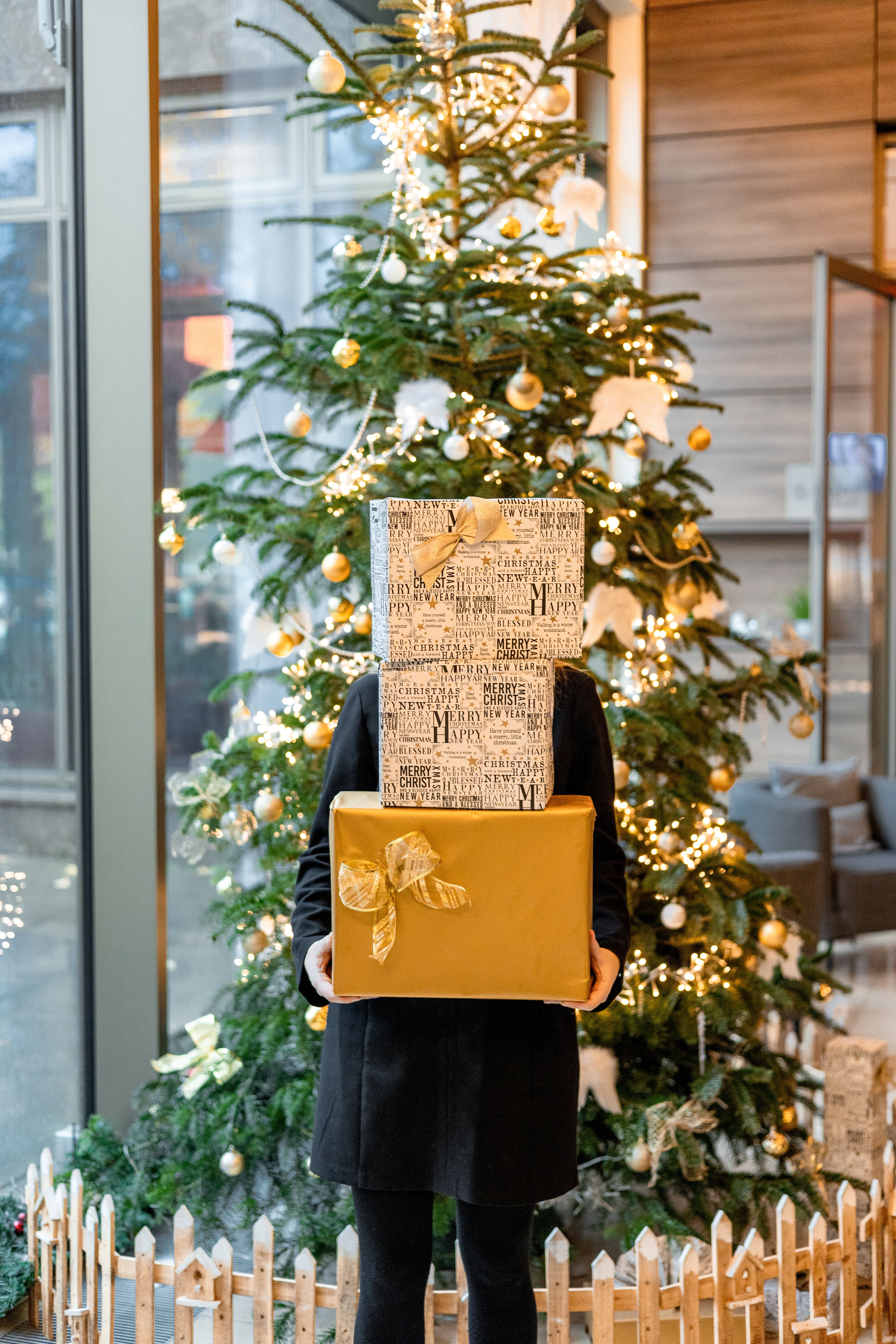 Person holding festive stacked Christmas presents near a lit Christmas tree.